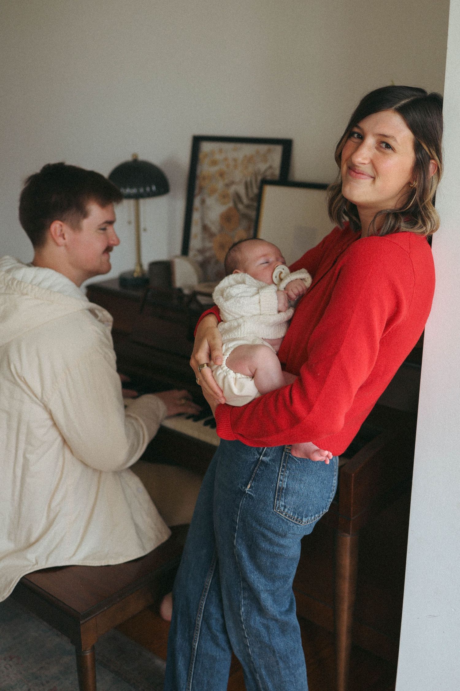 A woman in a red sweater holds a newborn baby while a man plays piano in a cozy living room.