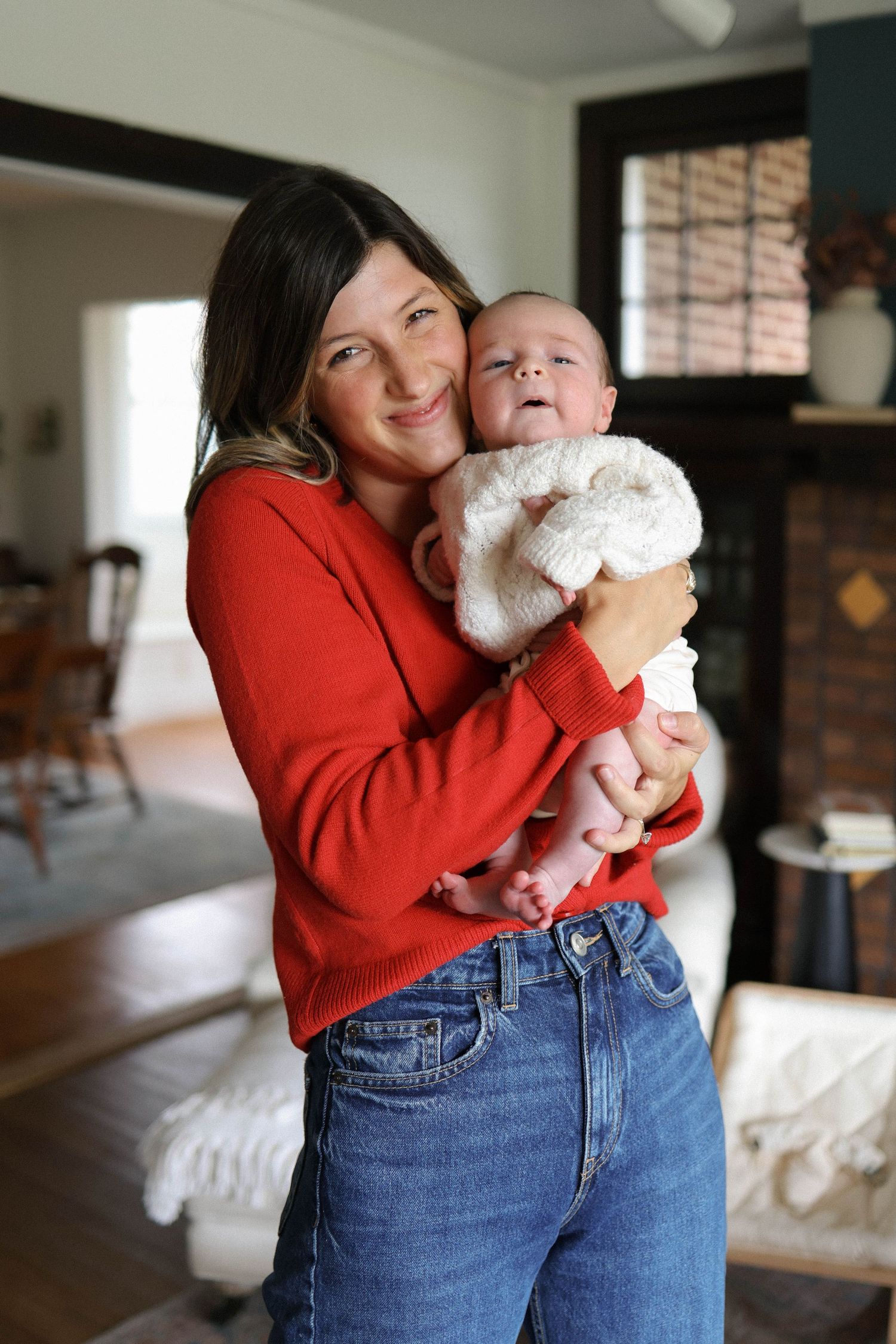 A smiling woman in a red sweater and jeans holds a newborn baby in a white knit outfit indoors.