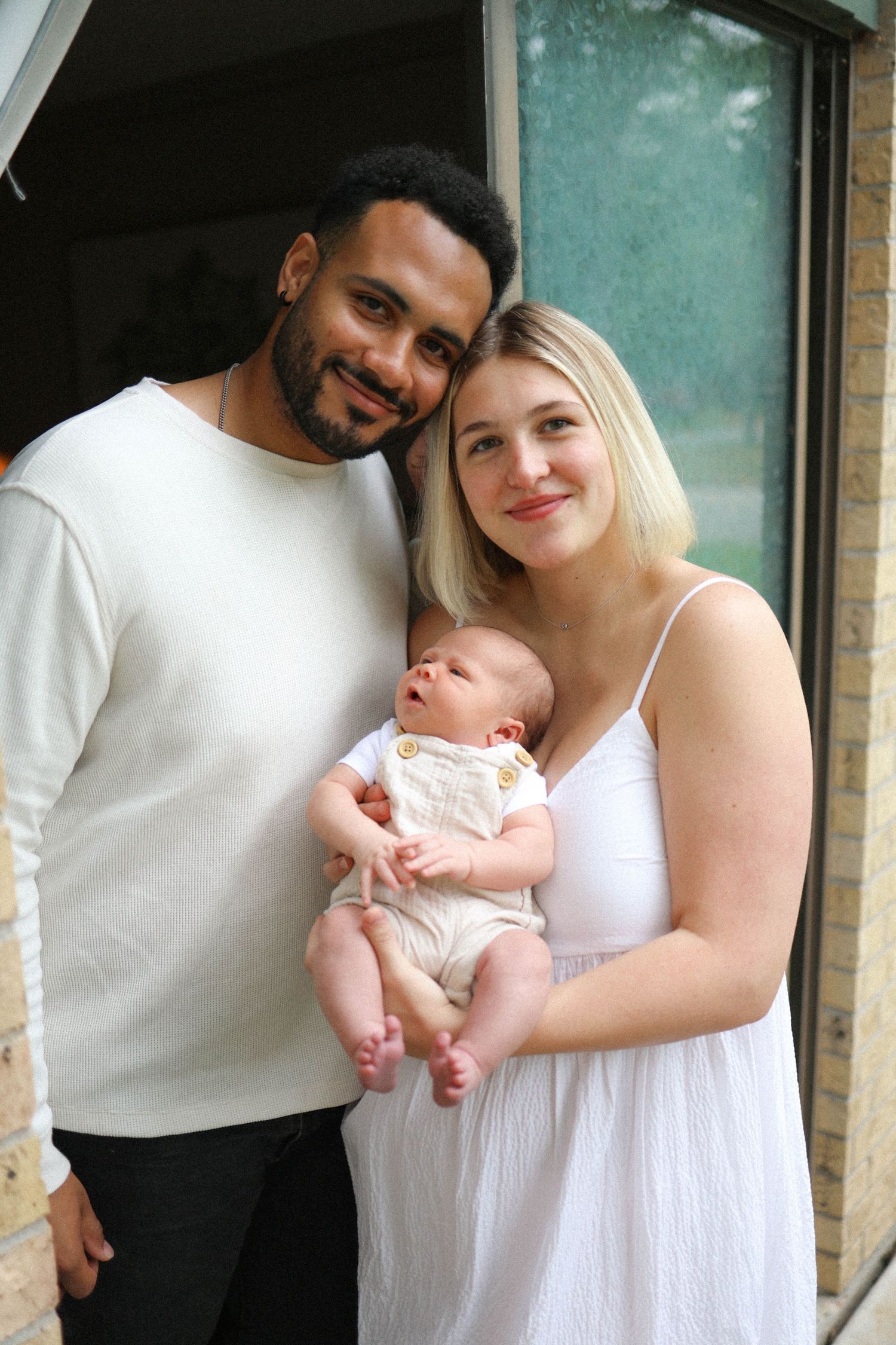 A couple with their newborn baby posing together against a brick wall exterior.