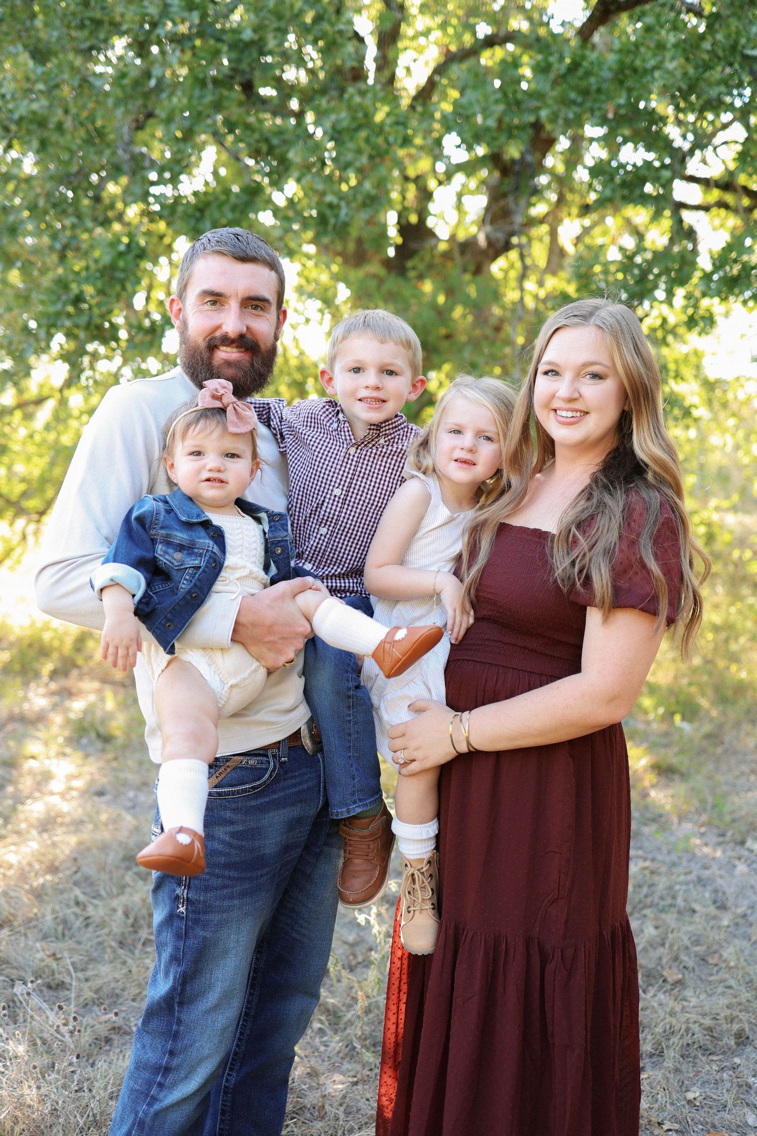 Smiling family of four with pregnant mother in burgundy dress poses outdoors under green trees for a portrait.
