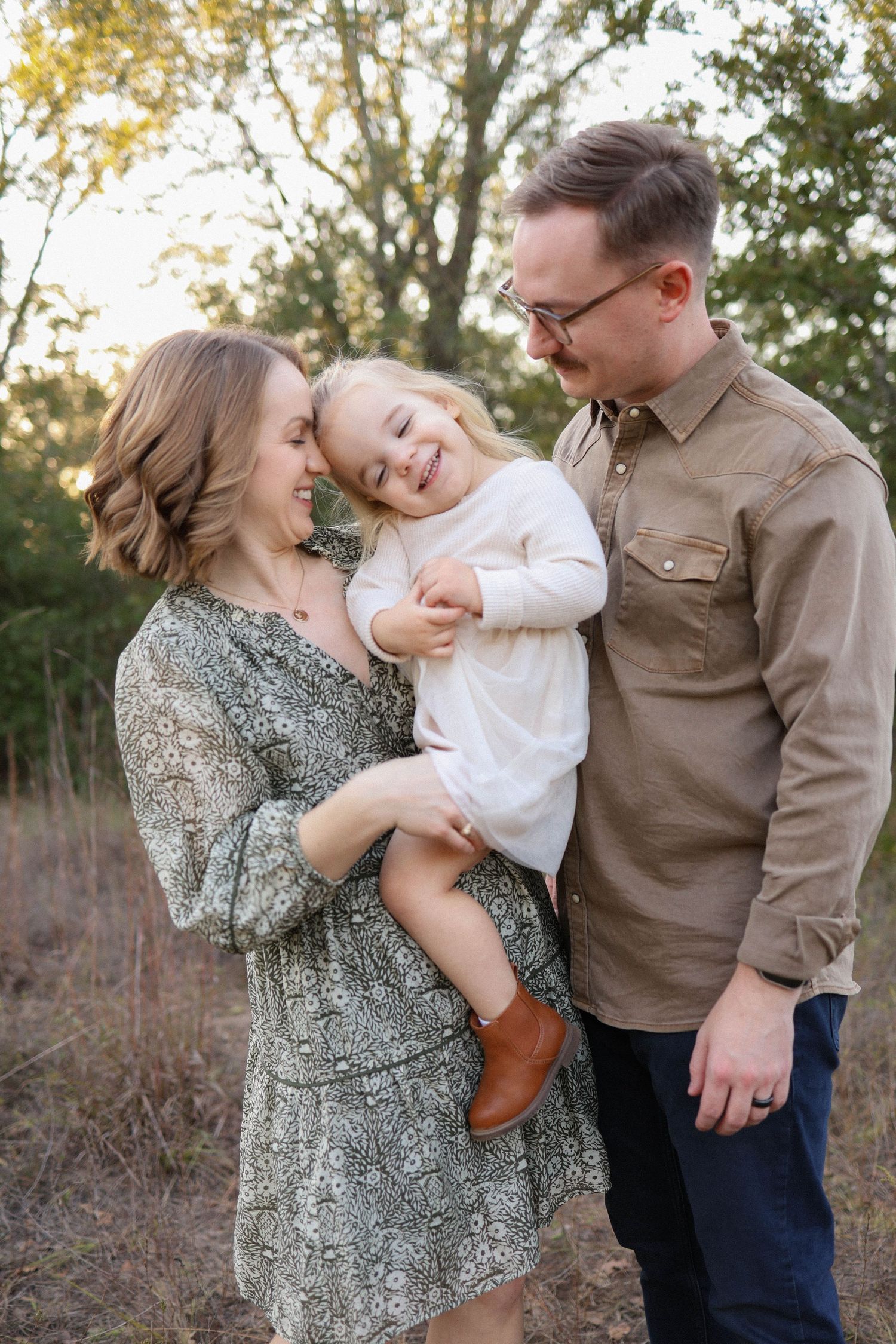 A happy young family with a toddler girl smiling outdoors in a wooded setting during a fall photo session.