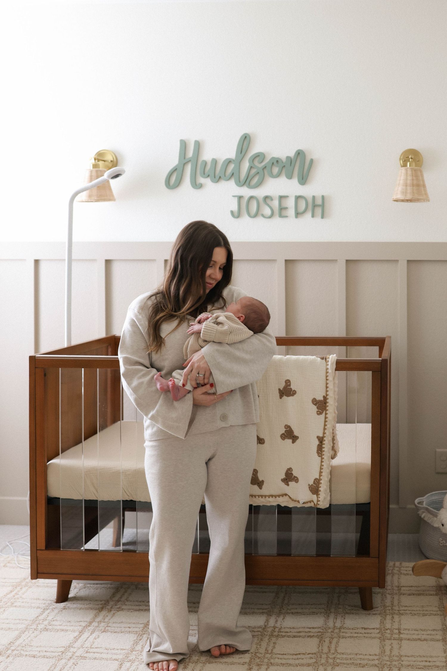 Mother holding newborn baby in nursery with Hudson Joseph name sign above wooden crib with plaid blanket.