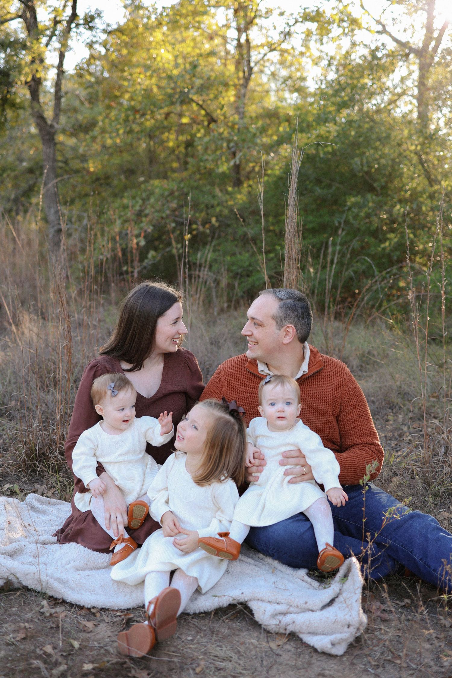 Family of five sitting on a blanket outdoors in autumn, parents smiling at each other with three young children in white outfits.