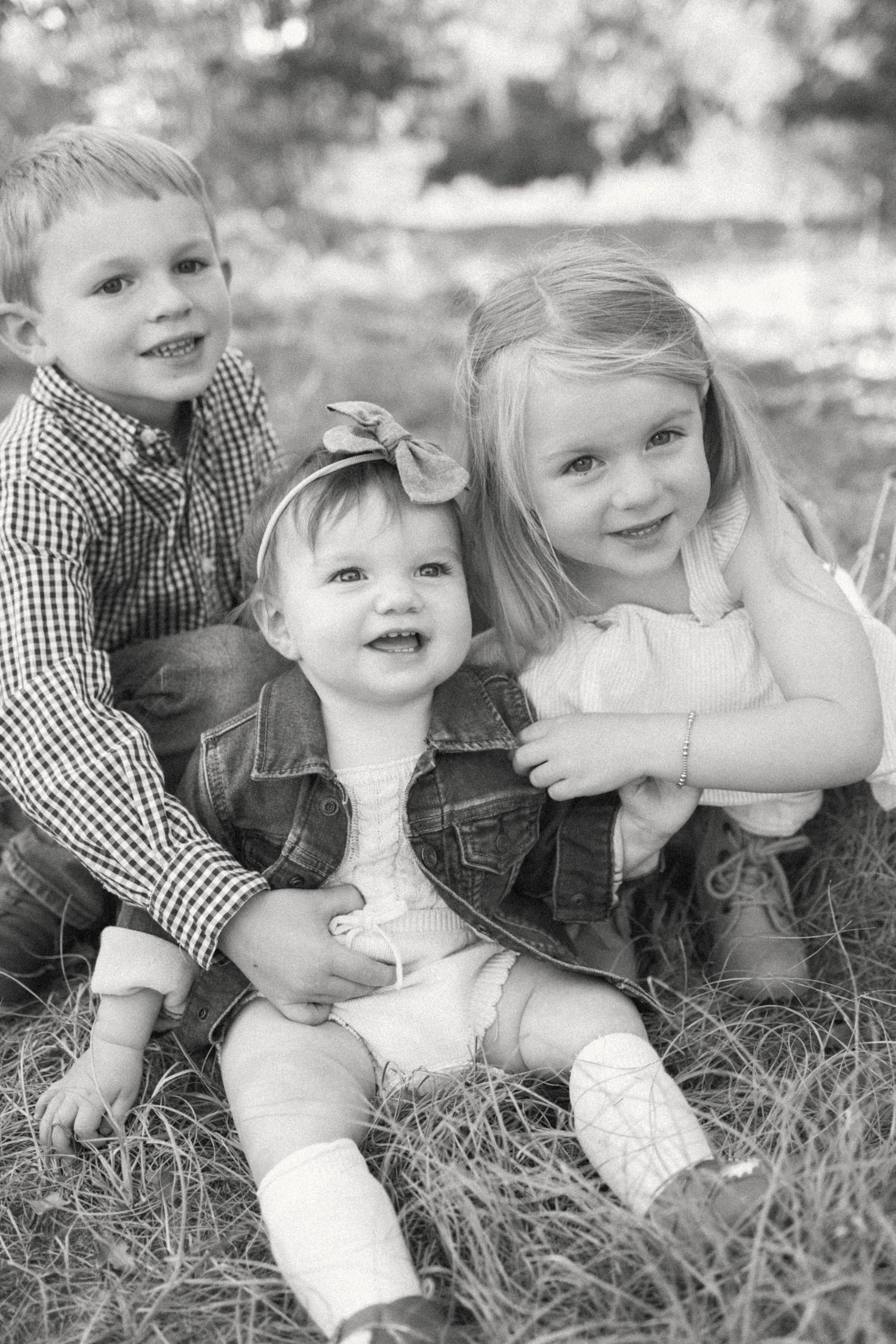Black and white portrait of three young siblings sitting together in grass, smiling warmly for a family photo.