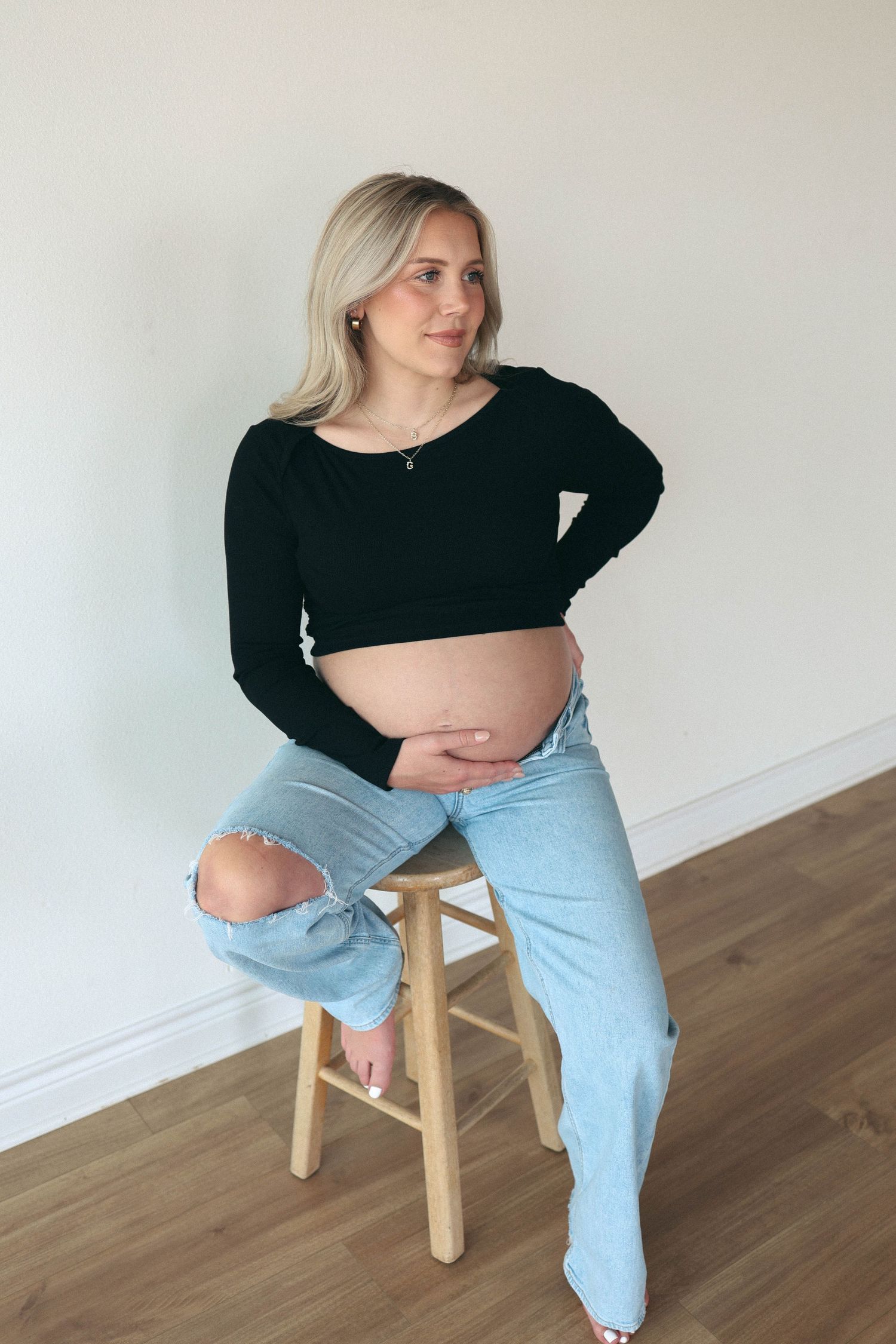 Pregnant woman in black crop top and ripped jeans sitting on wooden stool, cradling baby bump.