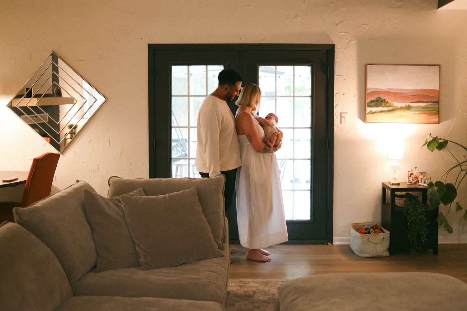 Couple sharing a tender moment holding newborn baby in a warmly lit living room with French doors.
