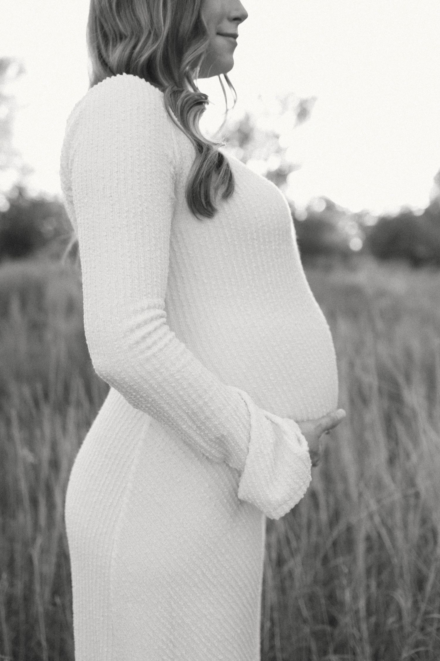 Black and white maternity photo of pregnant woman in white knit dress cradling baby bump in a field.