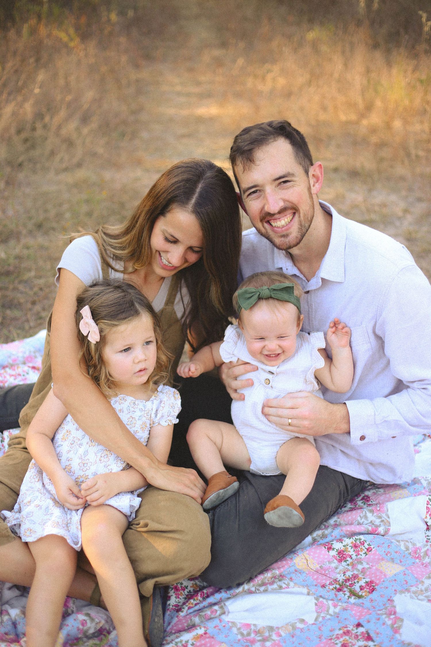 A family sits together on a floral blanket outdoors during a sunny portrait session.