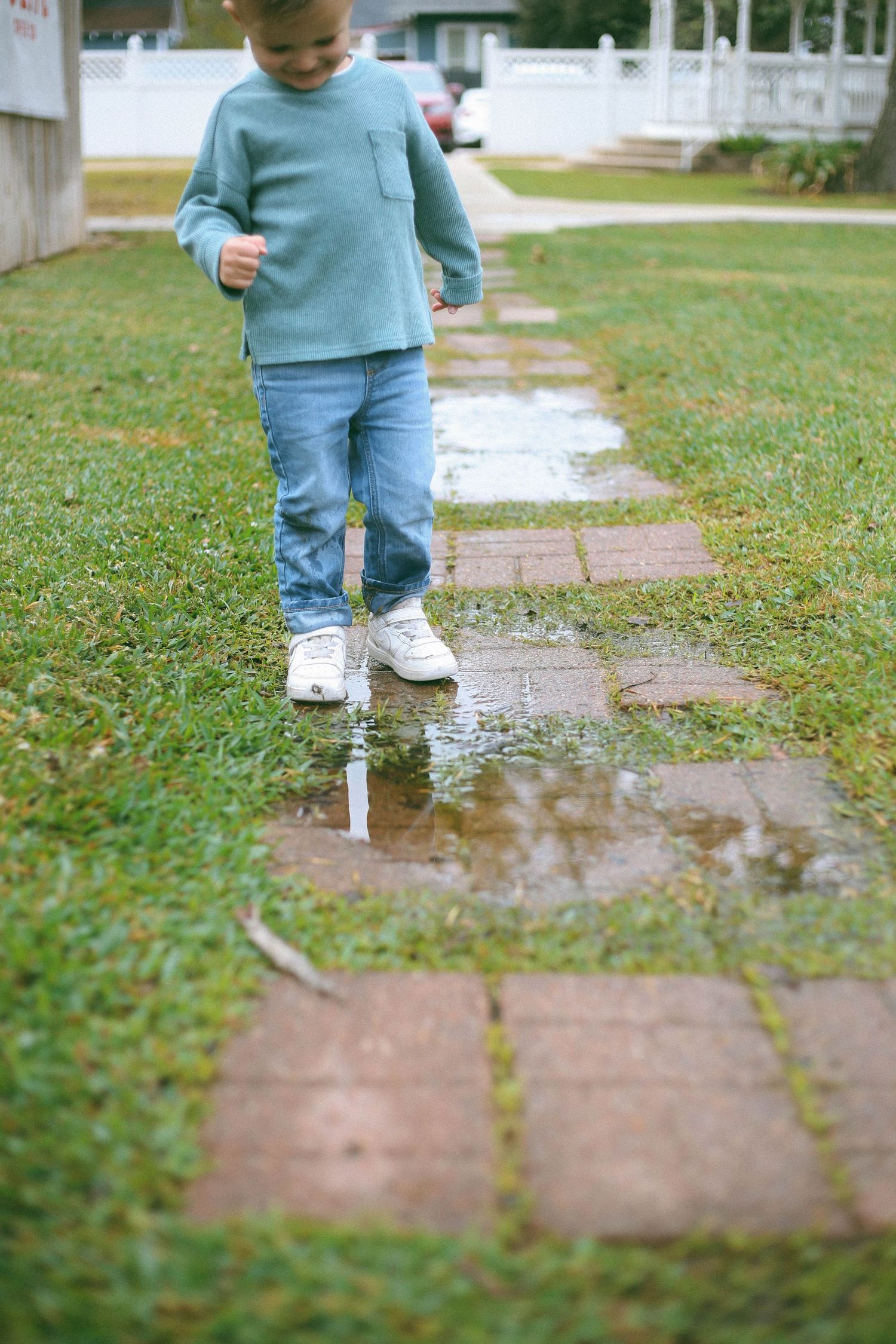 Child in blue outfit walking through puddles on brick pathway in garden setting.