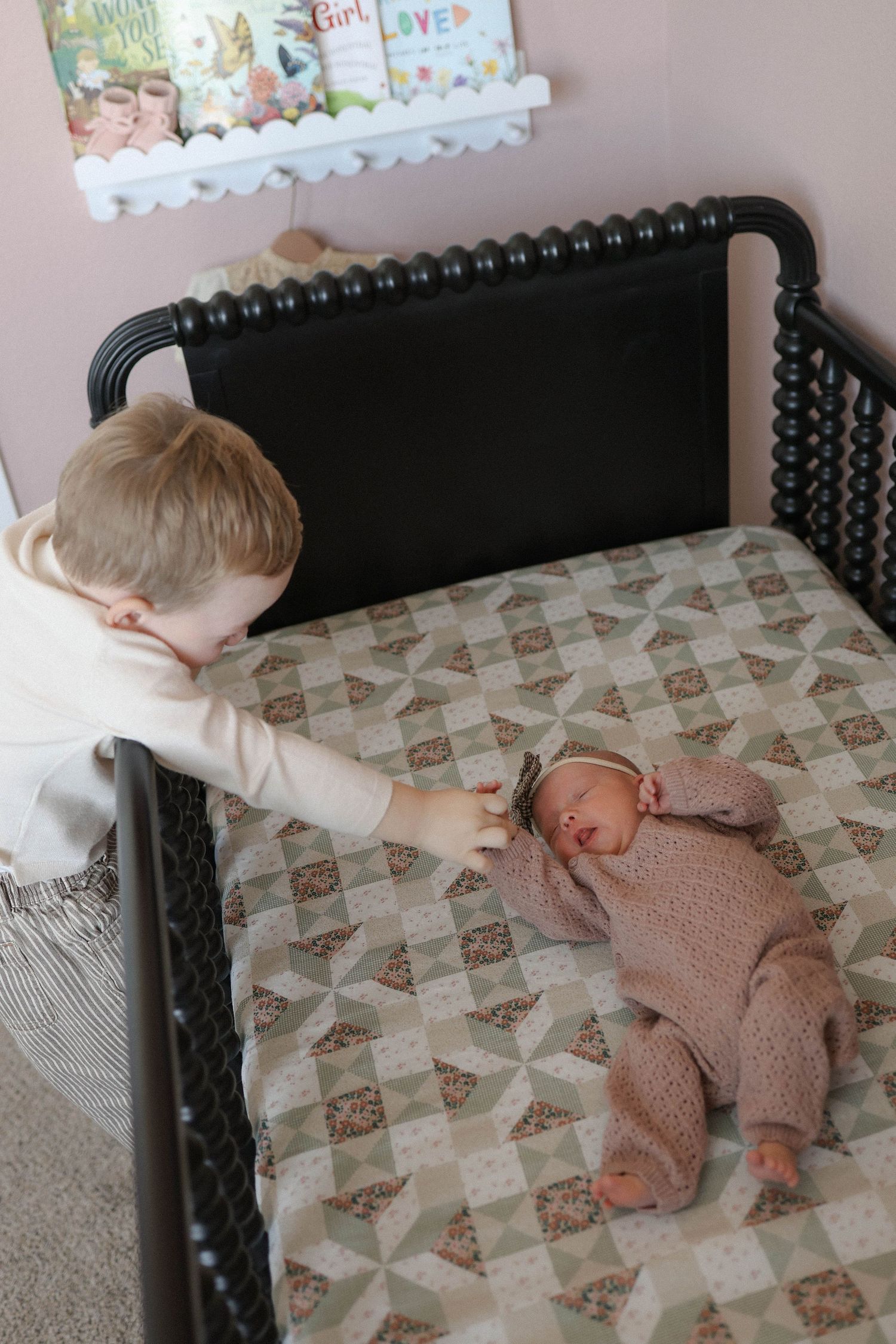 Toddler boy leaning over black jenny lind crib to touch newborn baby sibling lying on a patchwork quilt.