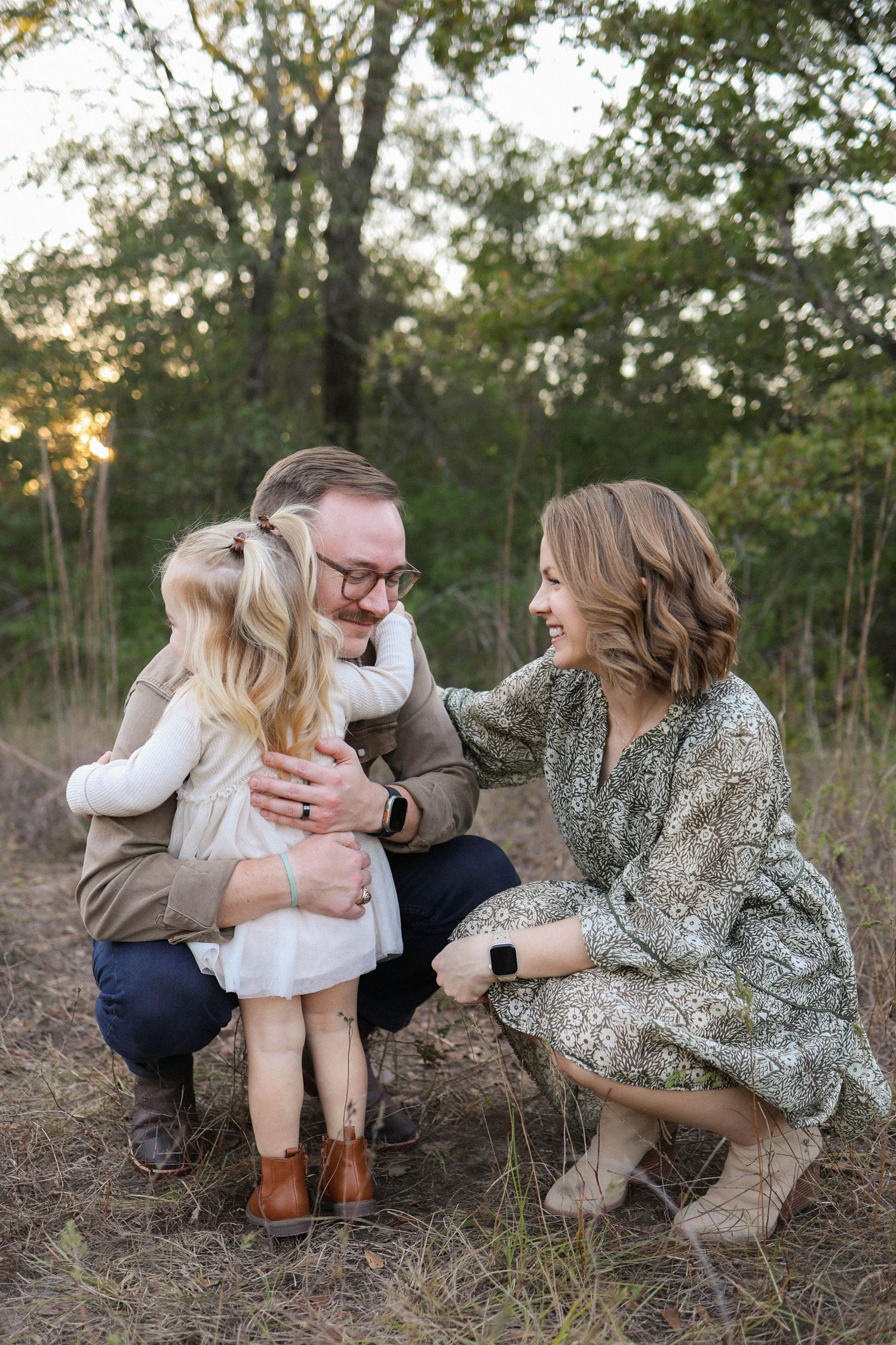 Family of three sharing a warm embrace outdoors at golden hour, parents kneeling with young blonde girl in white dress.