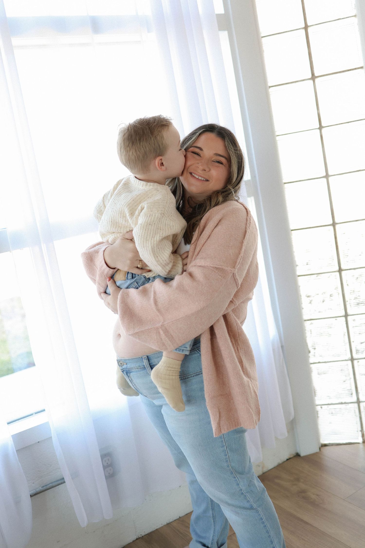A smiling woman in a pink sweater and jeans holds a baby who gives her a kiss on the cheek near bright windows.
