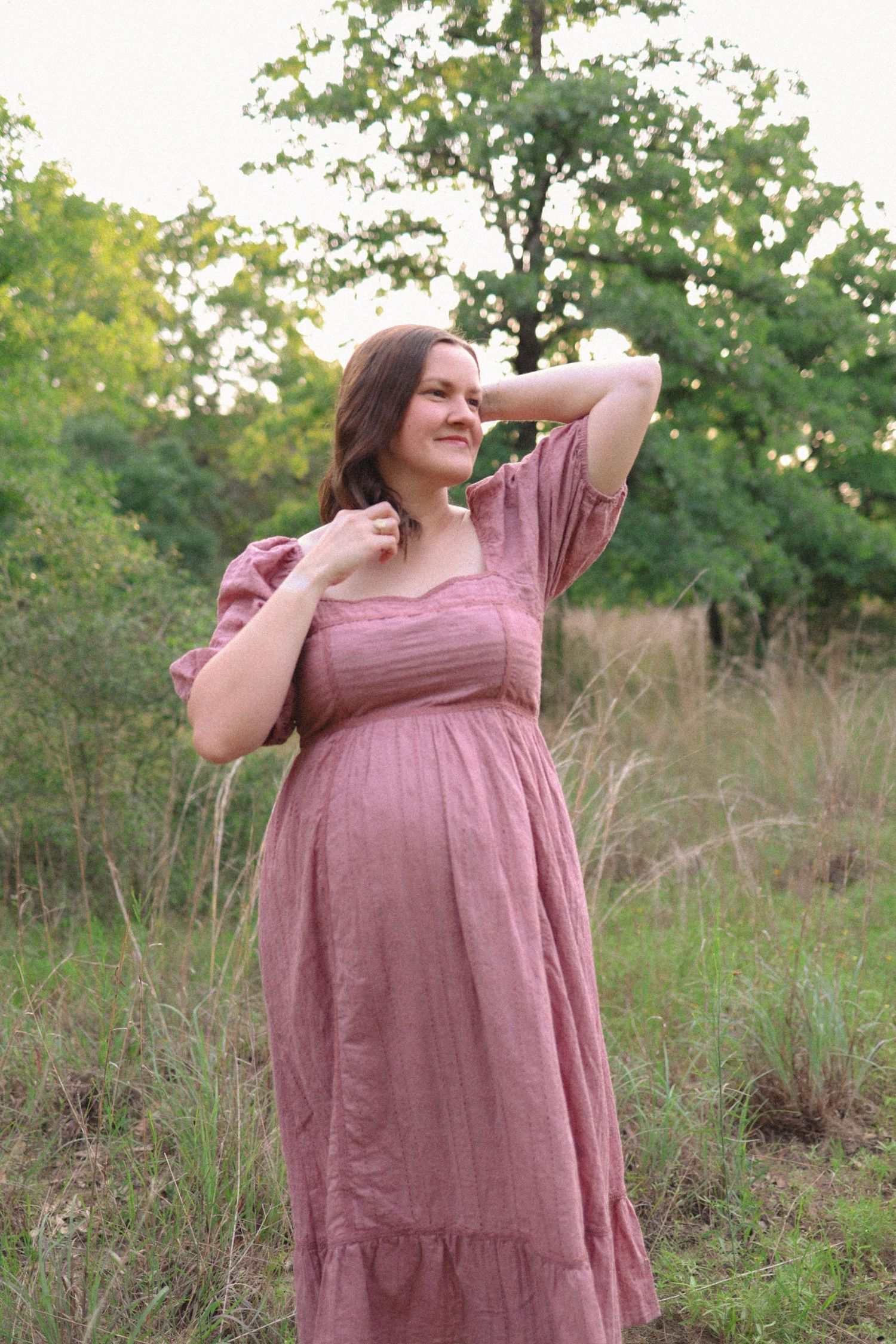A pregnant woman in a dusty rose dress poses gracefully in a sunlit field.