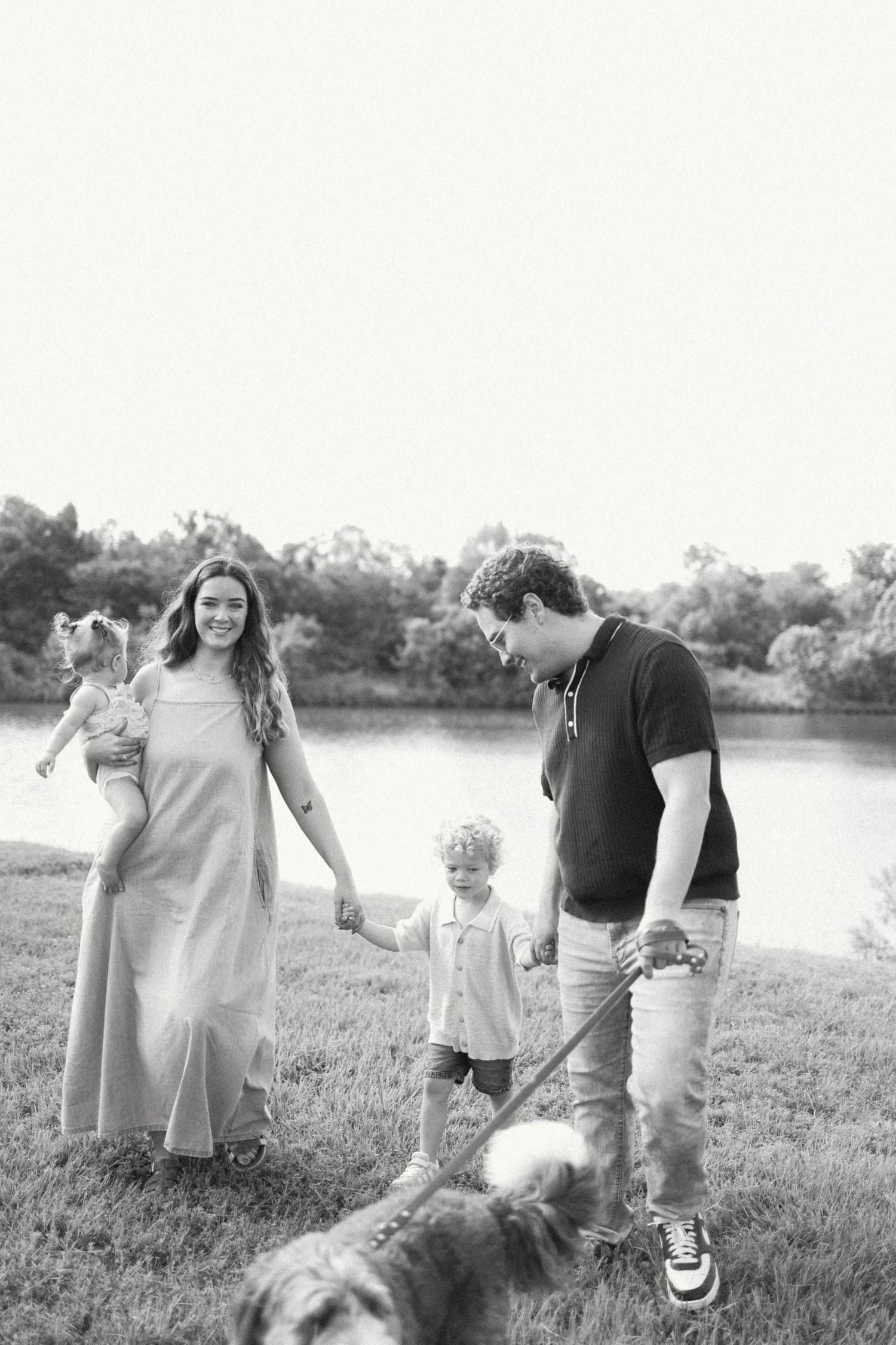 Black and white family photo with parents, two young children, and a dog walking by a lake outdoors.