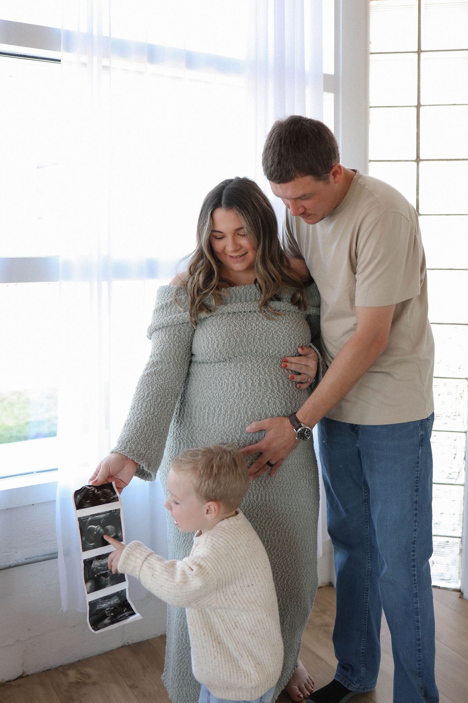 Pregnant woman with husband and young child holding ultrasound photos in bright home setting.