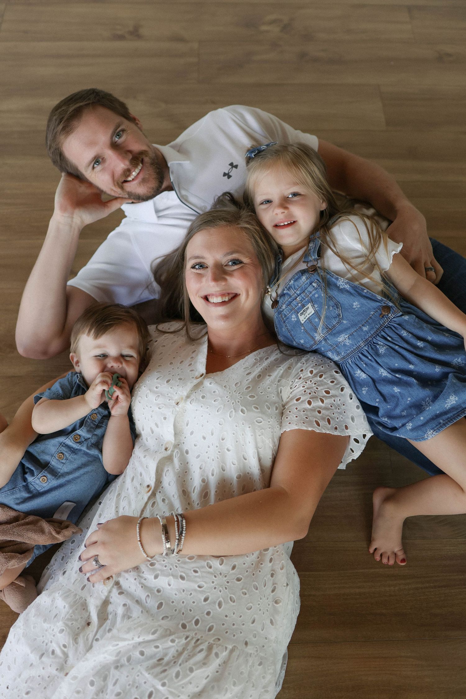 Happy family of four lying on wooden floor, parents smiling with two young daughters in denim outfits.