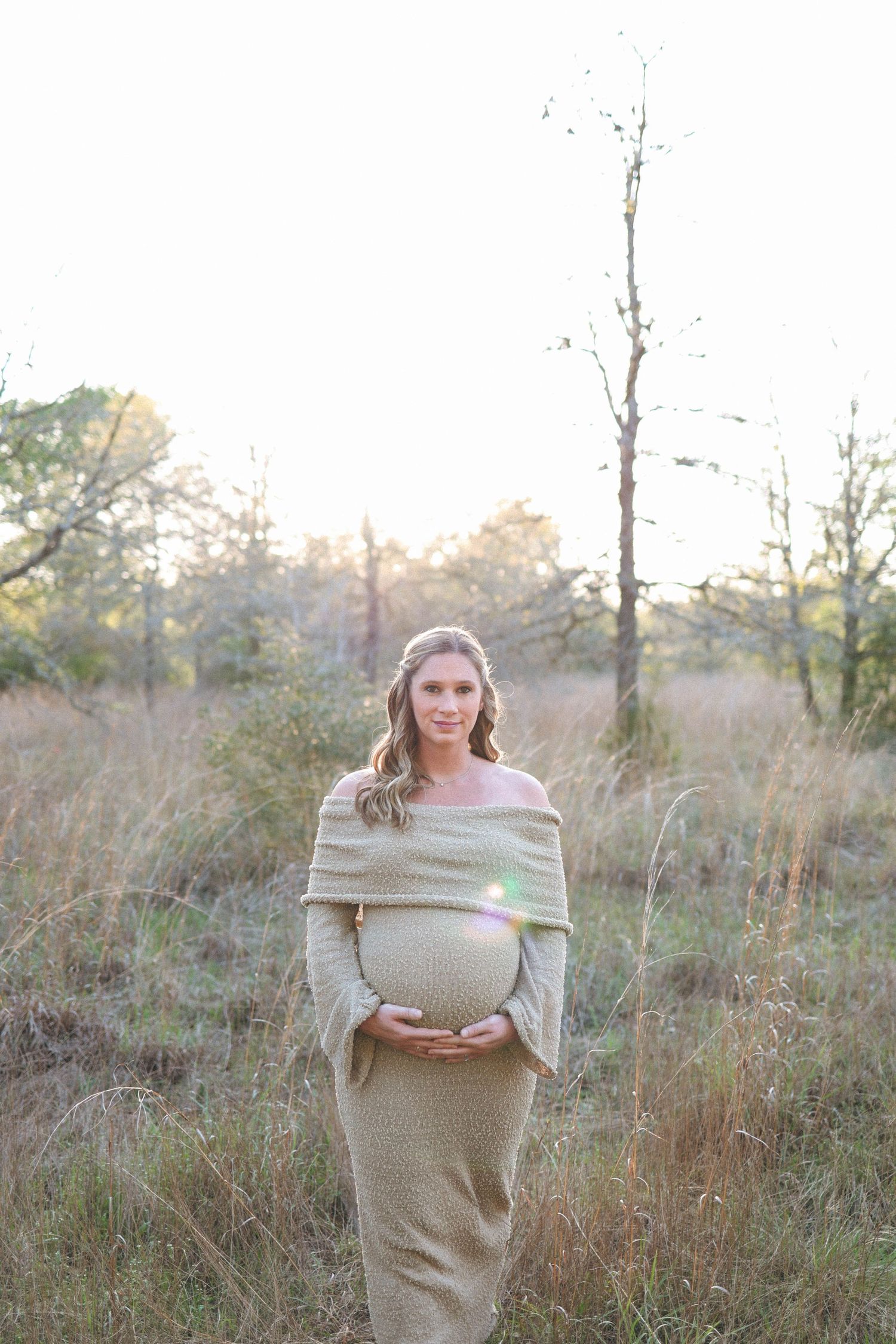 Expecting mom in beige dress stands in sunlit field during golden hour maternity photoshoot.