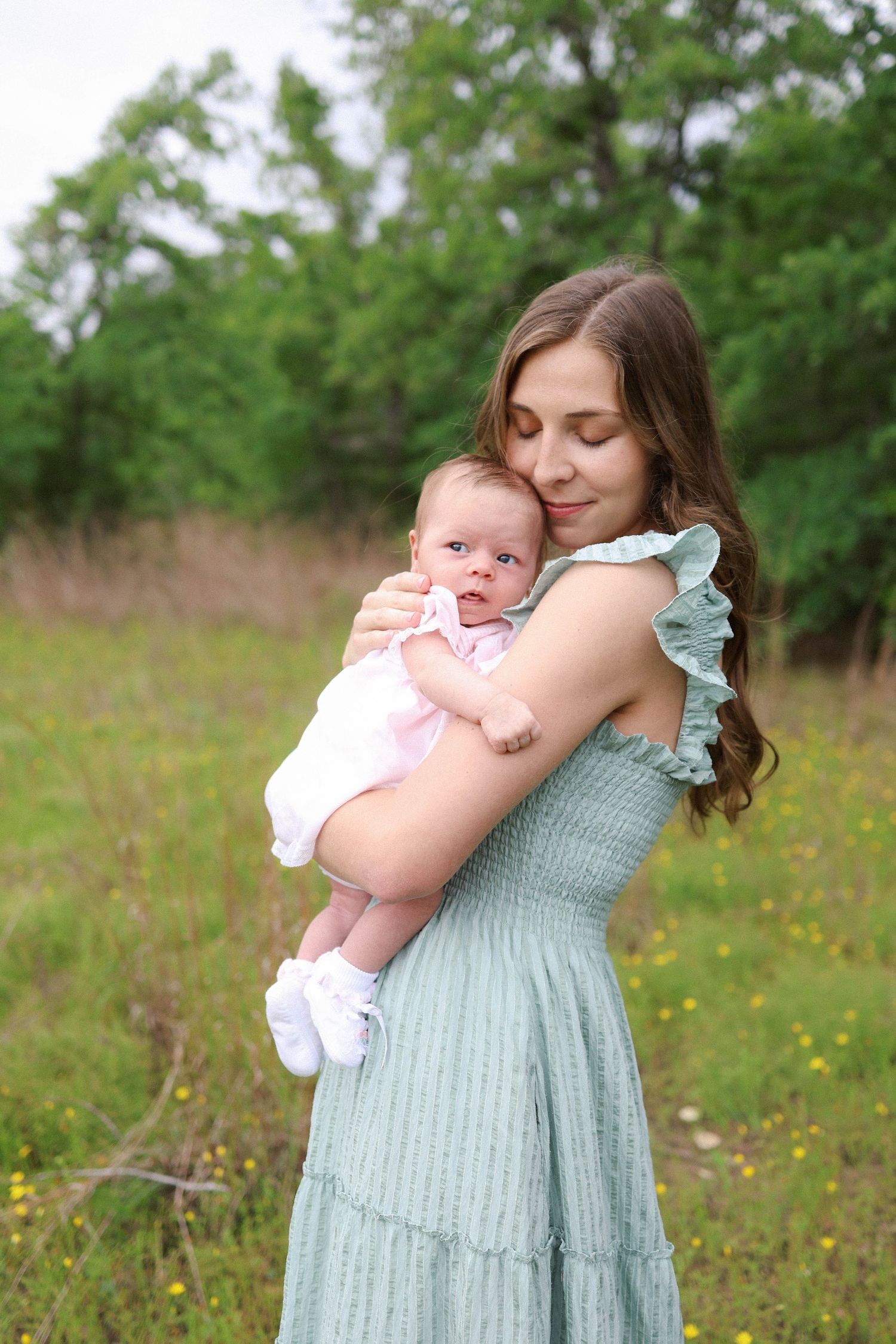 Young mother in teal dress tenderly holds newborn baby outdoors in green meadow with wildflowers.