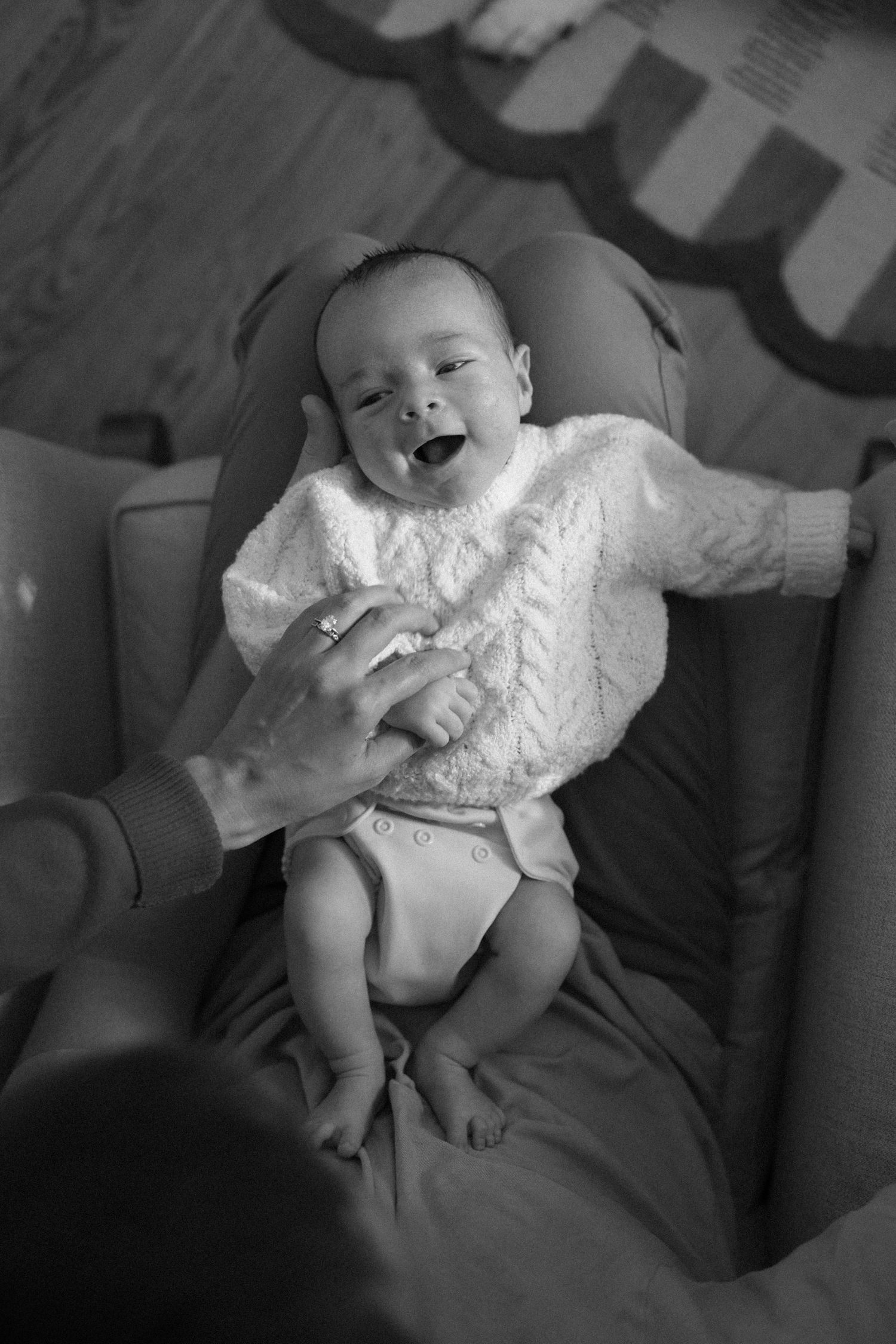 Black and white photo of a smiling newborn baby in a white knit sweater, held by an adult hand in a baby seat.