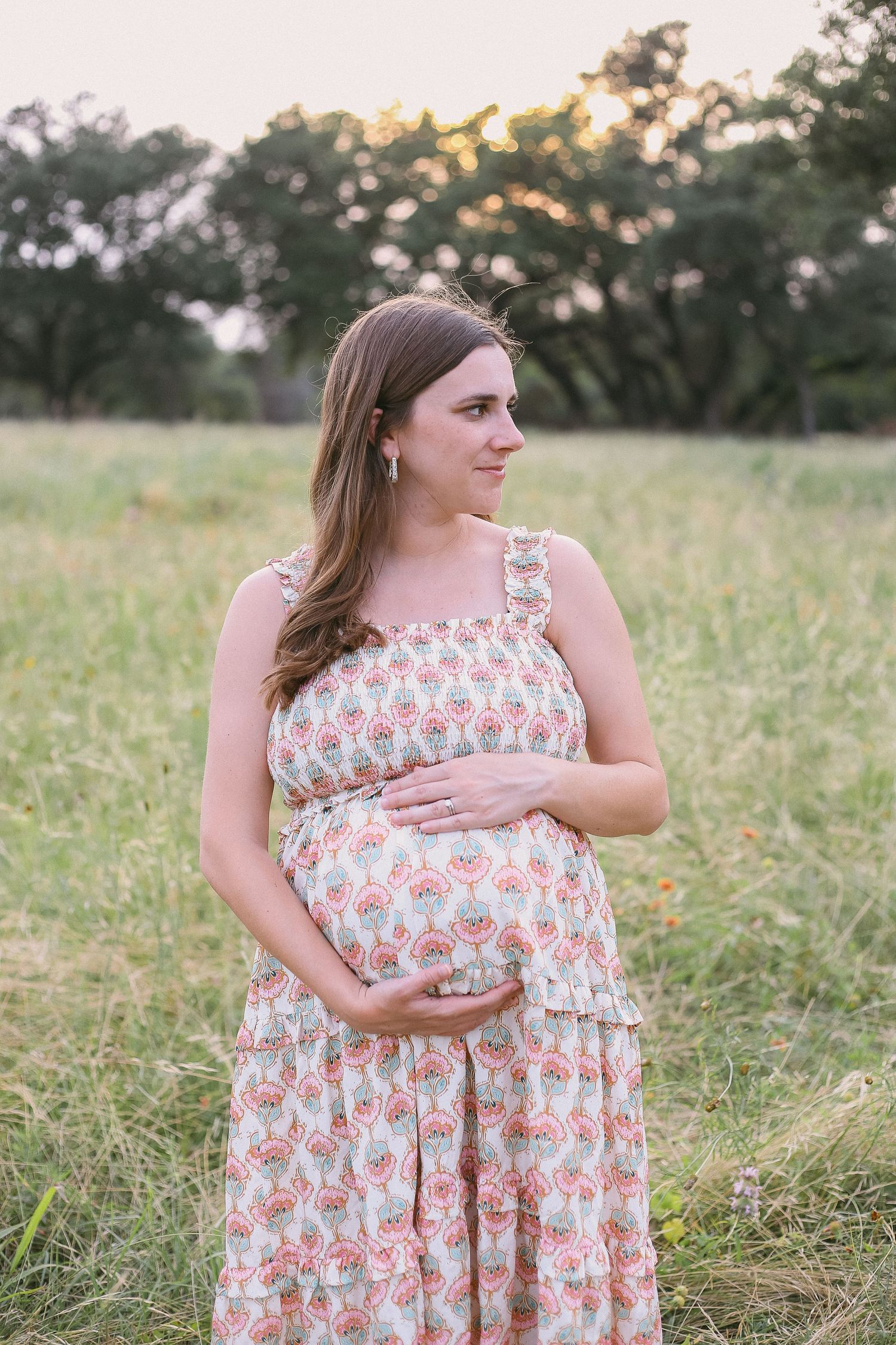 A serene maternity portrait of a woman in a floral dress standing in a golden field at sunset.
