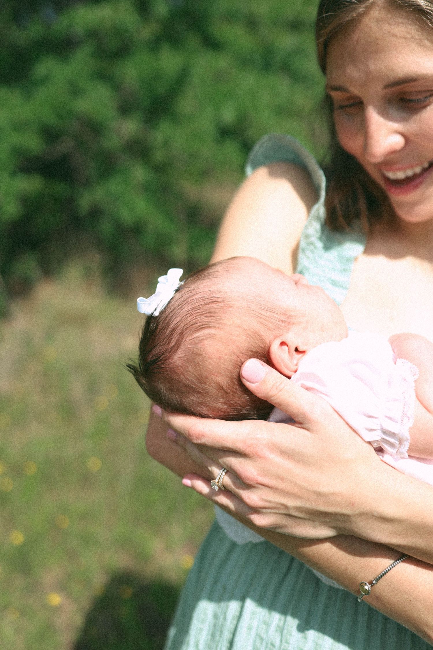 A smiling mother cradles her newborn baby in pink outdoors, with lush green trees in the background.