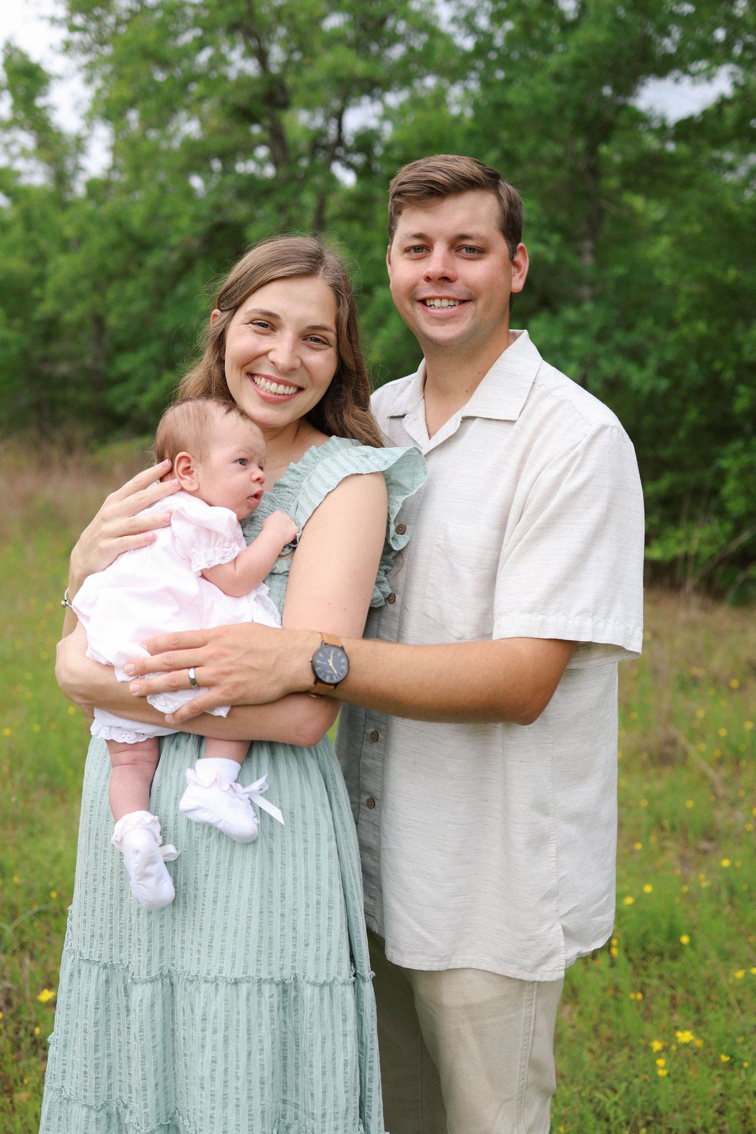 Happy young couple smiling outdoors holding their newborn baby girl in a green meadow setting.