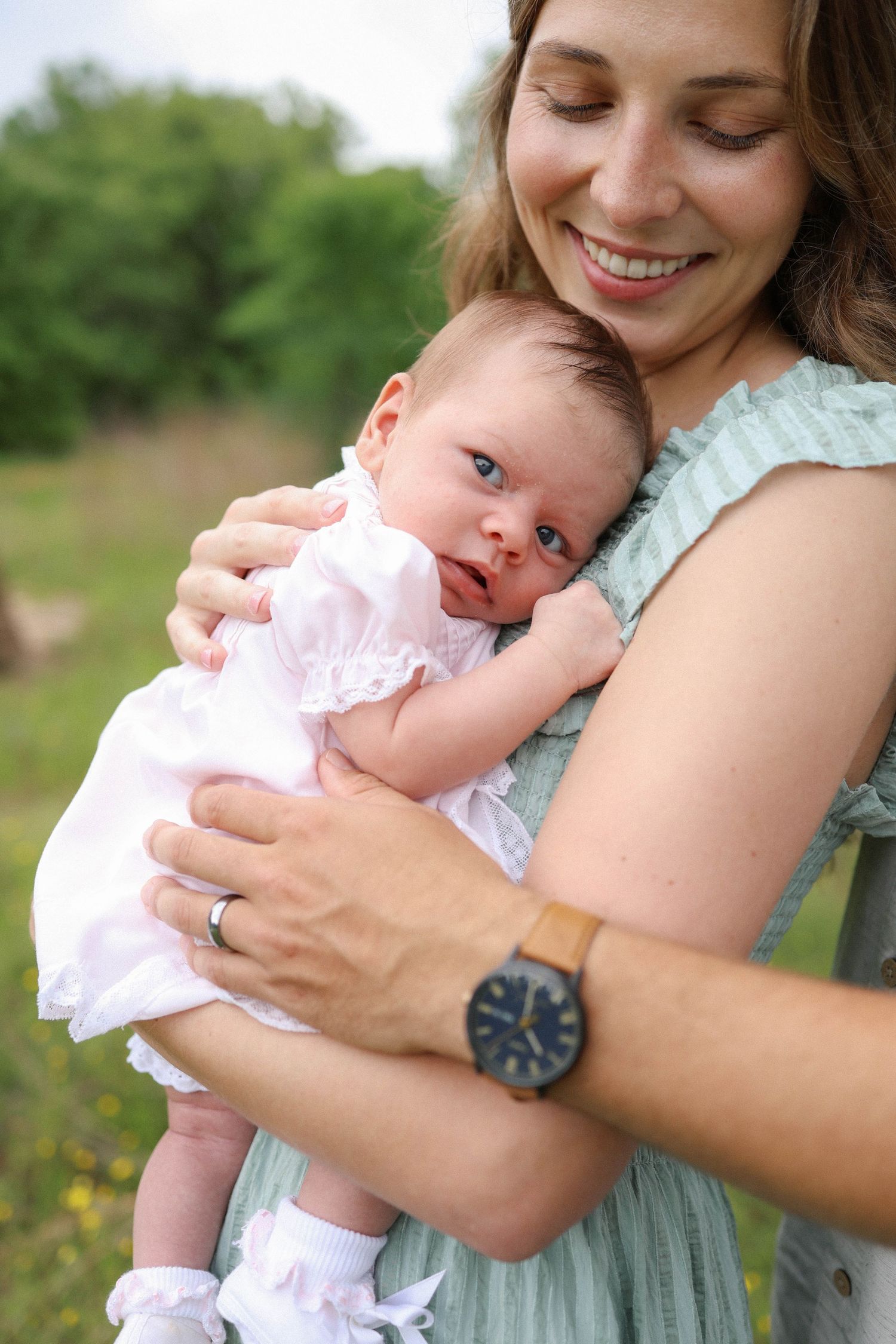 Smiling mother in mint dress tenderly holds her newborn baby in white dress outdoors in green nature setting.