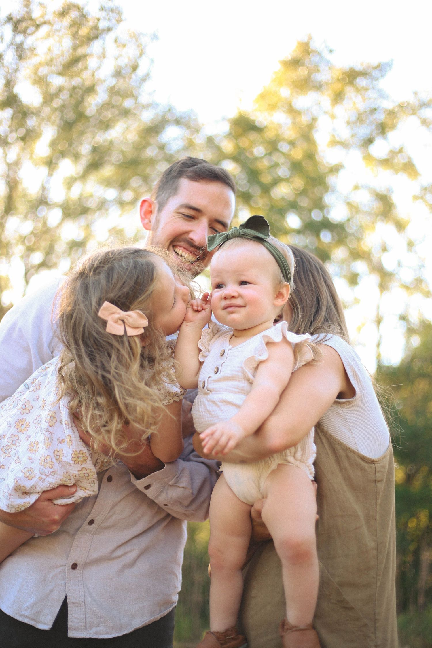 Playful family moment captured outdoors with parents kissing baby's cheeks.