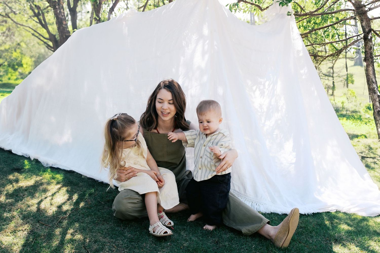A mother sits on grass with two young children in front of a white teepee tent outdoors.