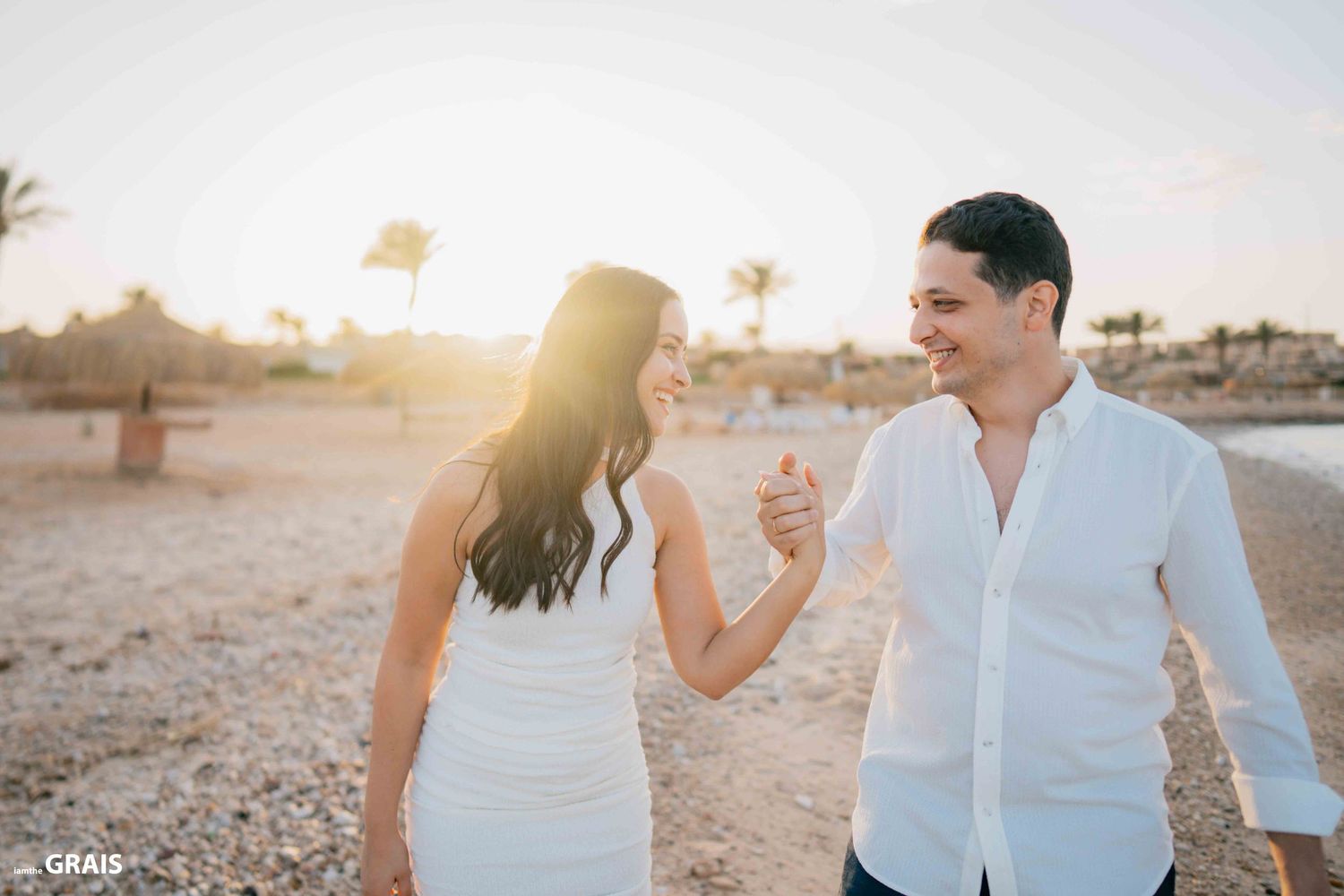 A couple in white outfits walks along the beach at sunset.