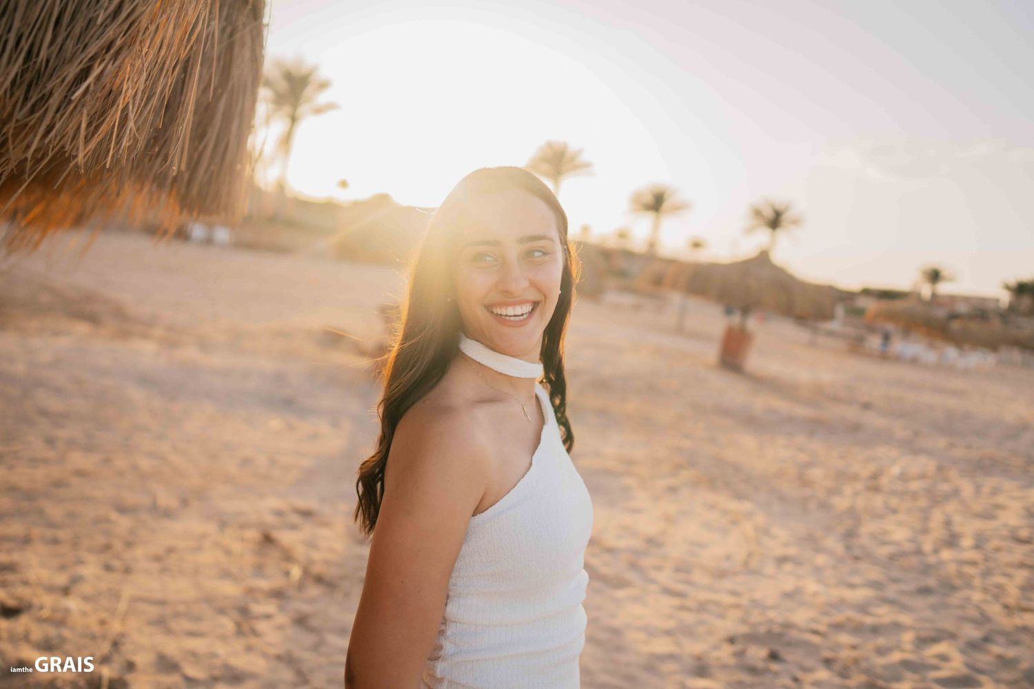 A woman in a white dress smiles brightly on a beach during golden hour.