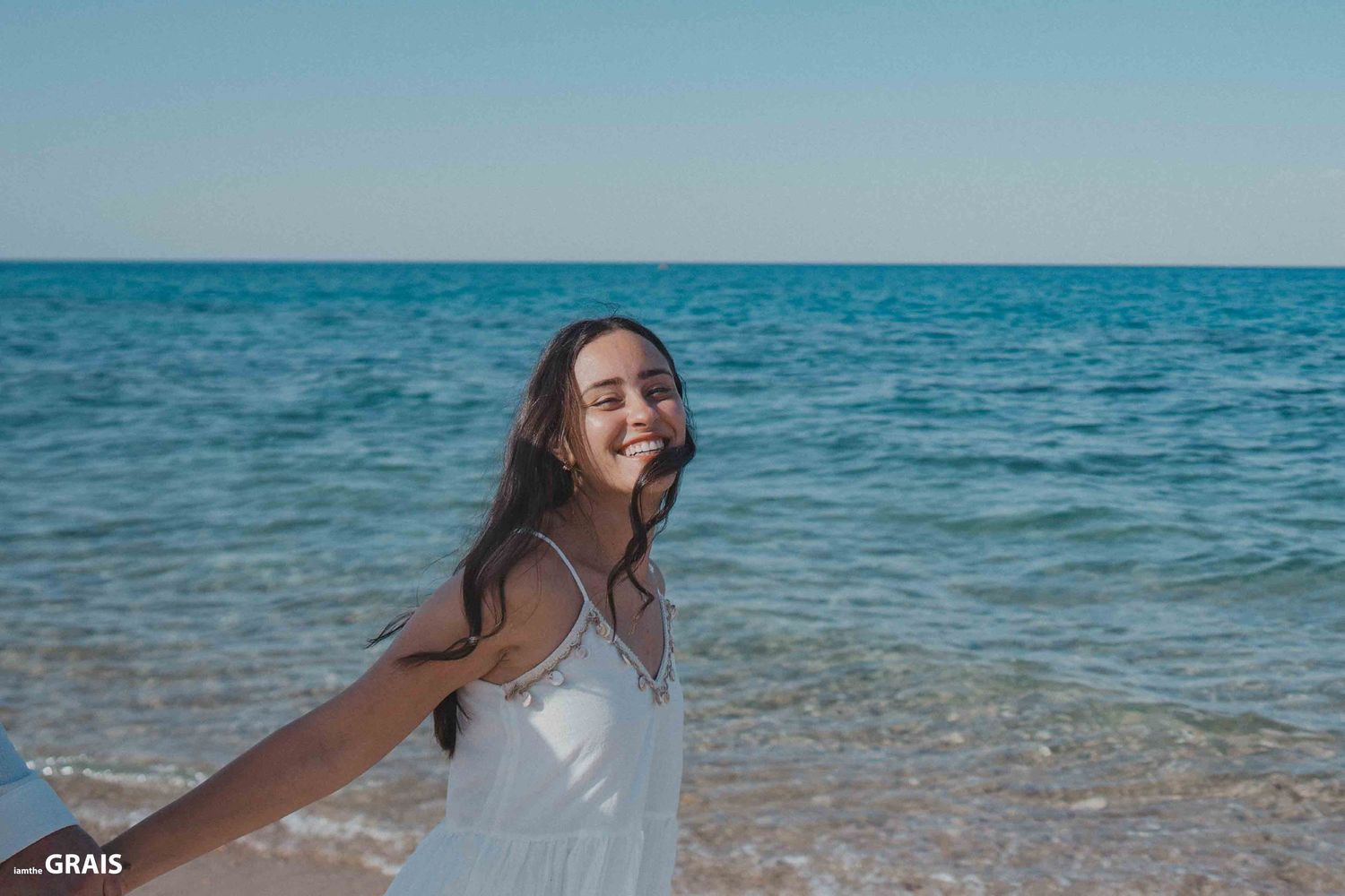 Woman in flowing dress poses freely against blue ocean backdrop.