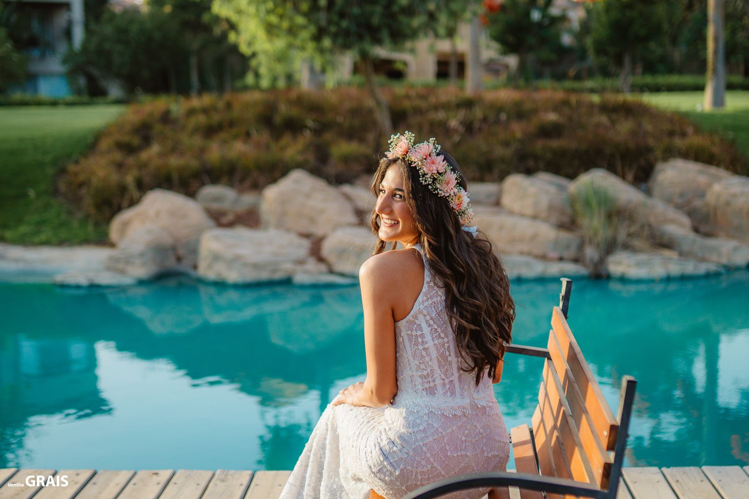Bride in lace dress sits by luxurious pool surrounded by rocks and greenery.