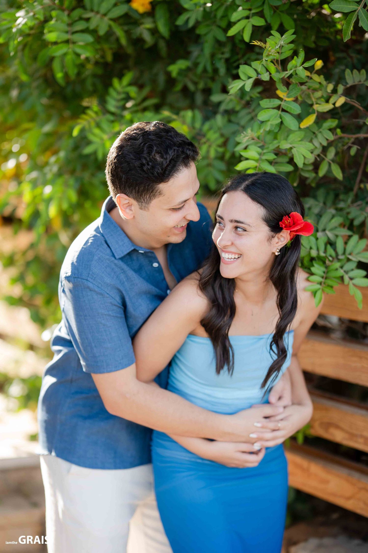 A couple embraces in a garden setting with the woman wearing a light blue dress and a red flower in her hair.