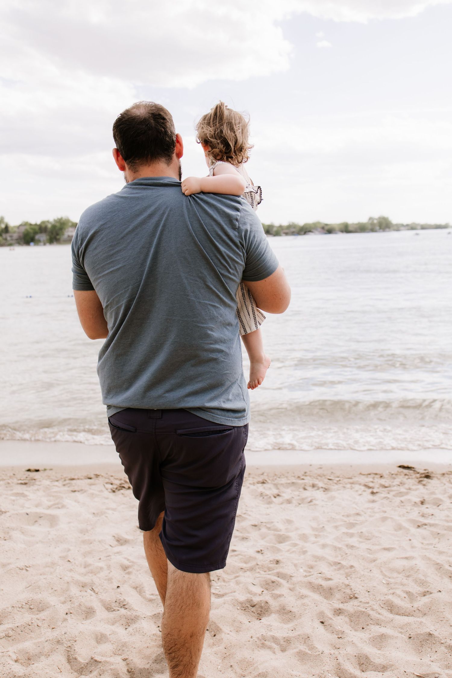 A father carrying his young child on the sandy beach, both facing the calm water under a cloudy sky.