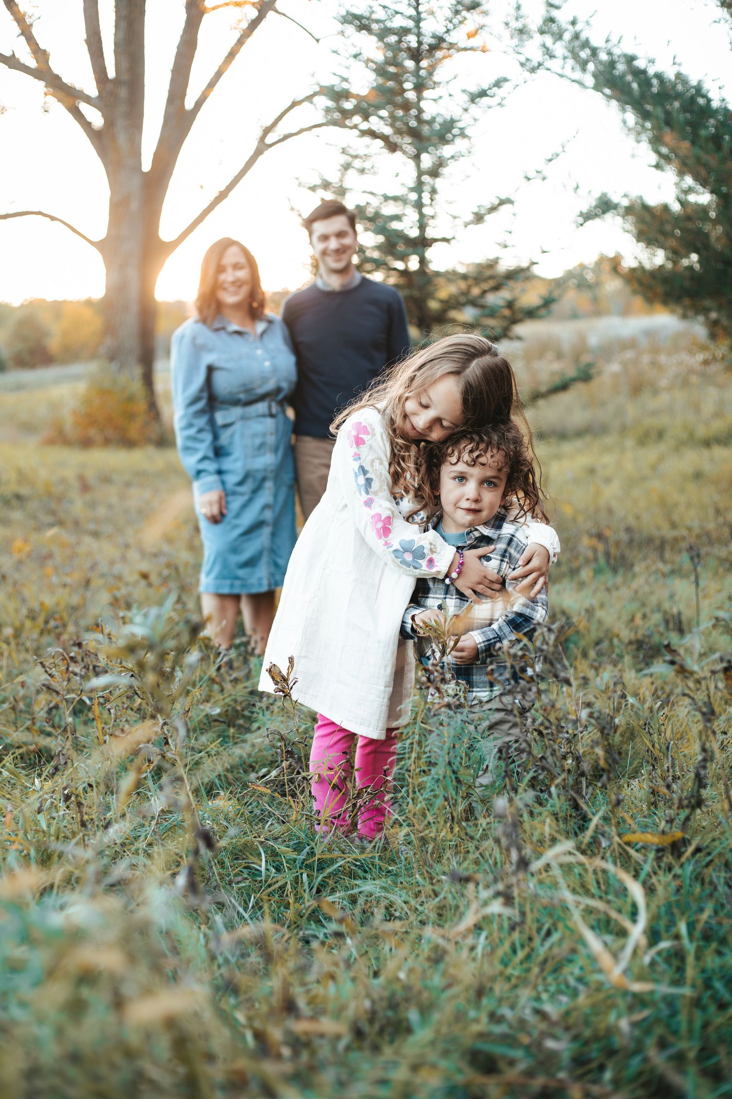 A young girl hugs her sibling in a sunny autumn field while their parents smile in the background, taken by Lakeville family photographer