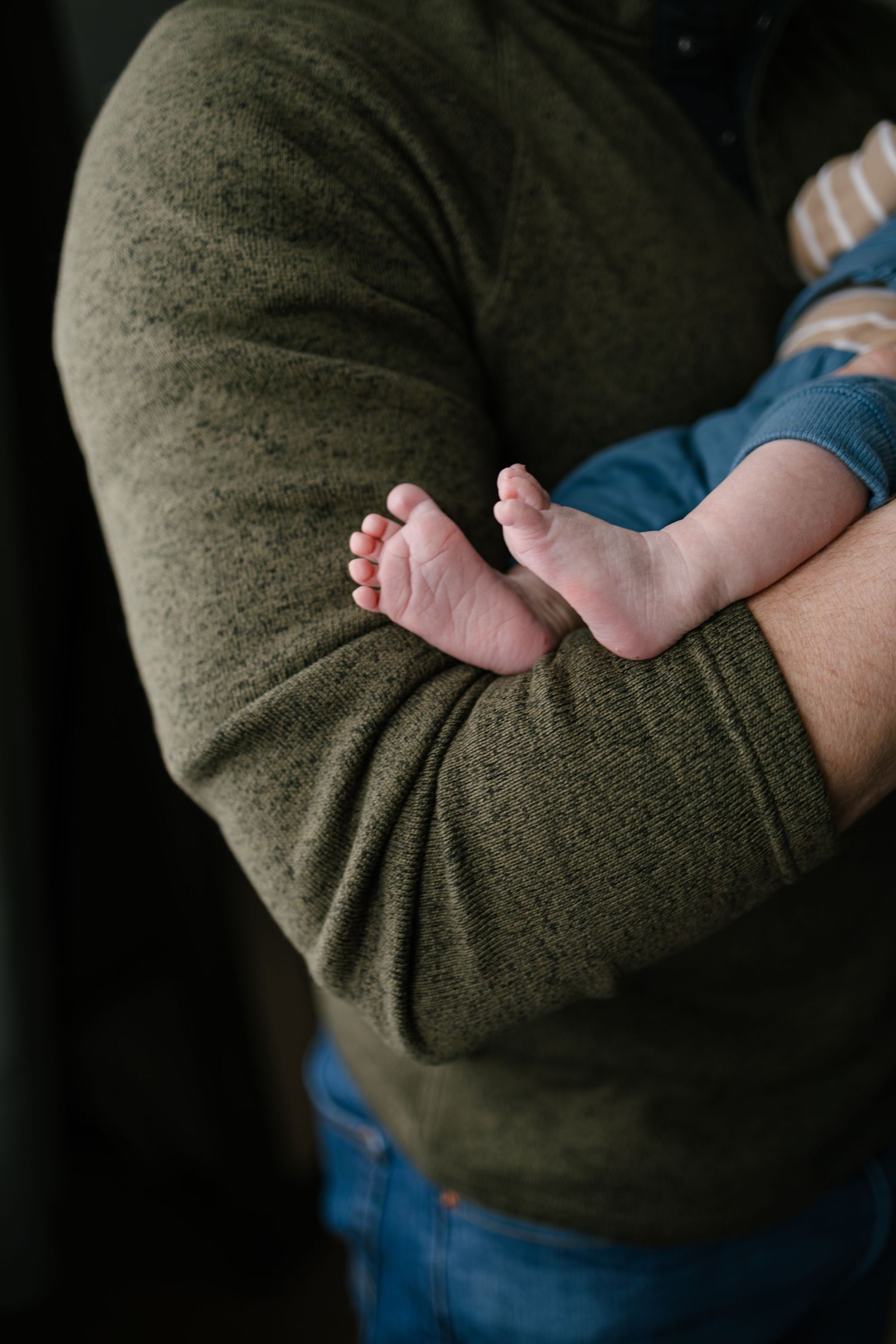 A close-up photo showing tiny baby feet resting against a dark green sweater sleeve, taken by Woodbury newborn photographer.