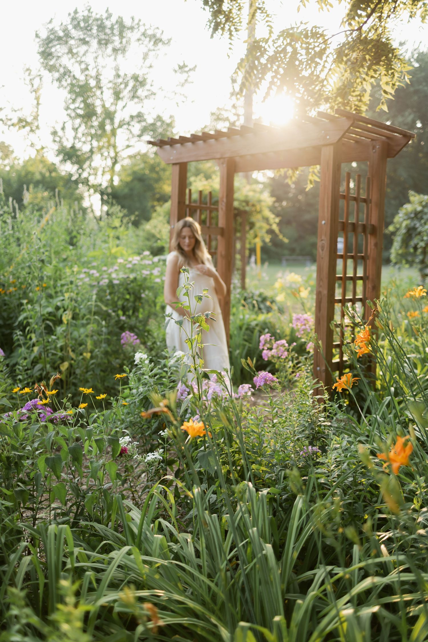 Woman in white dress walking through a lush flower garden at golden hour near a wooden arbor.