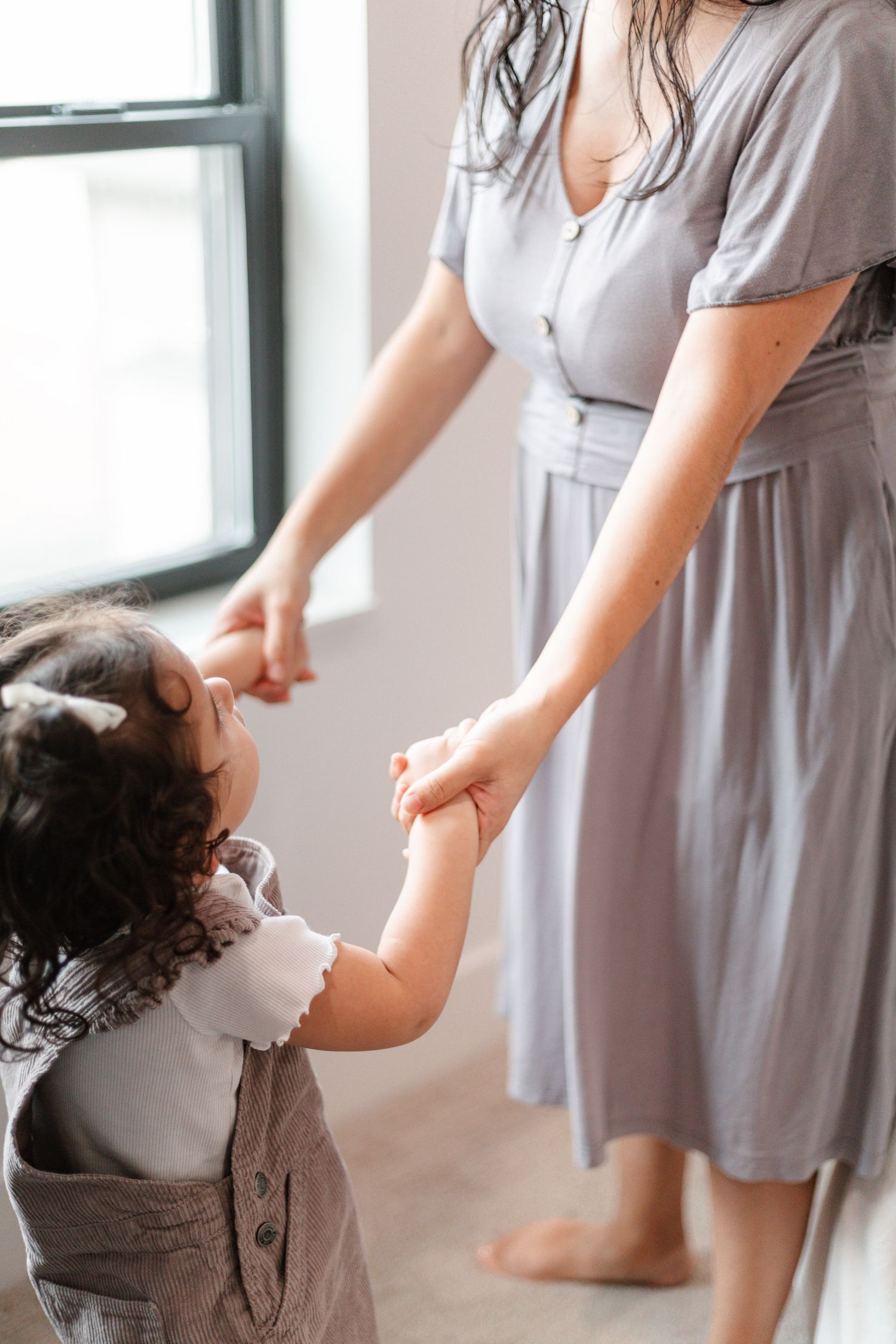 A mother in a lavender dress holds hands with her curly-haired toddler daughter near a bright window indoors in Minneapolis
