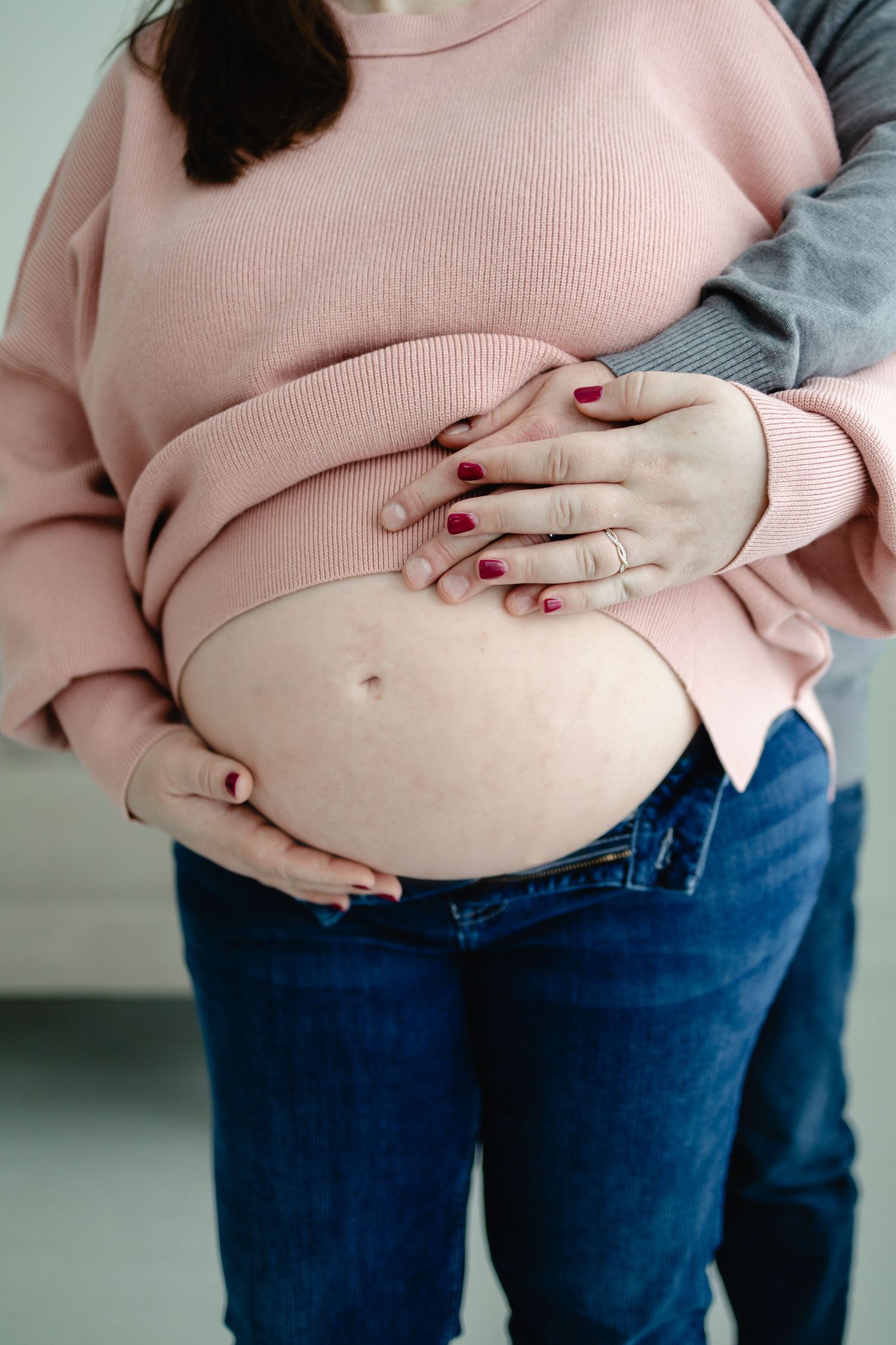 Pregnant woman in pink sweater and jeans cradling baby bump, partner embracing from behind captured by Woodbury photographer