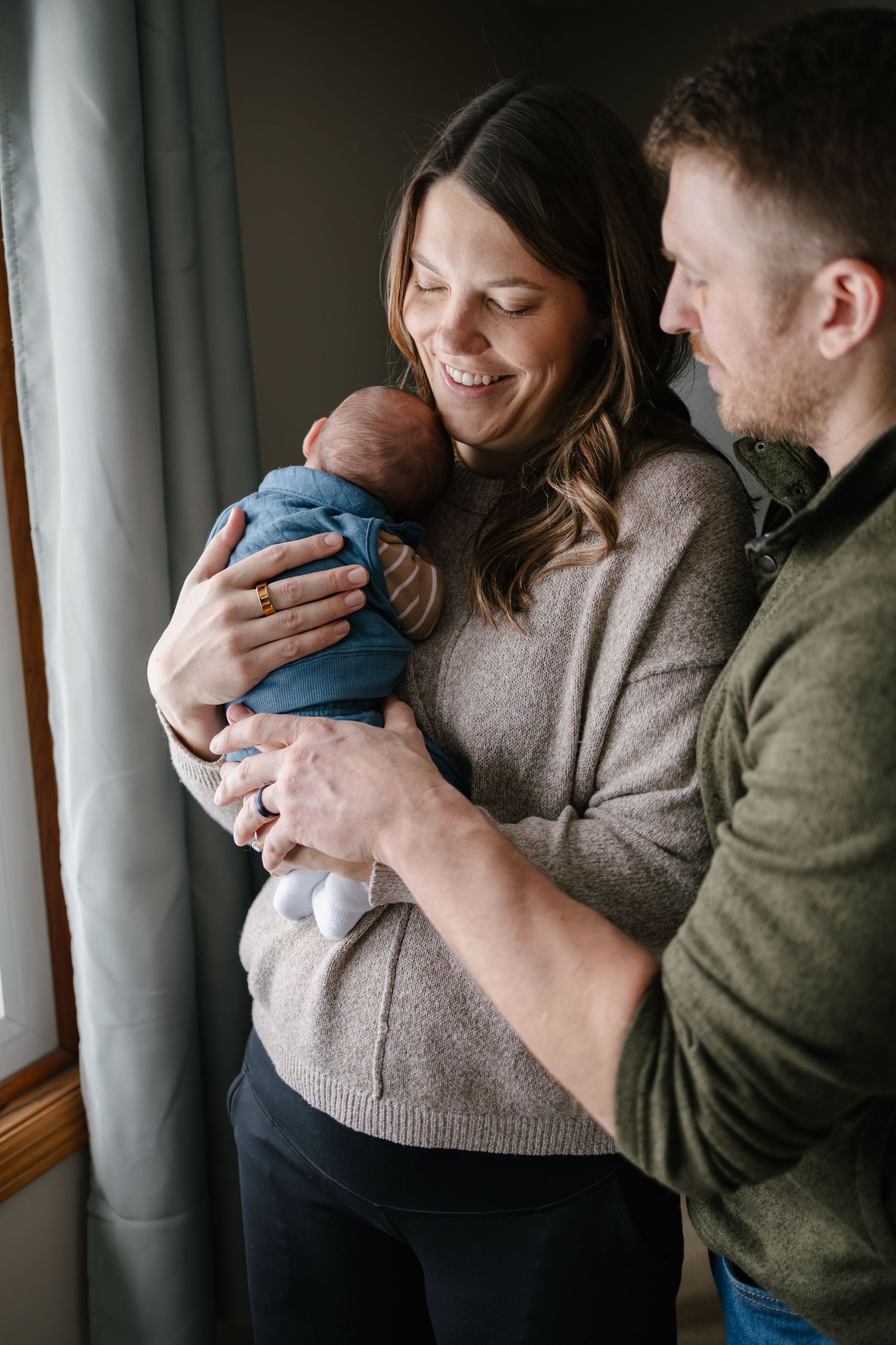 A couple stands by a window sharing a tender moment with their newborn baby wrapped in blue, taken by Woodbury newborn photographer.