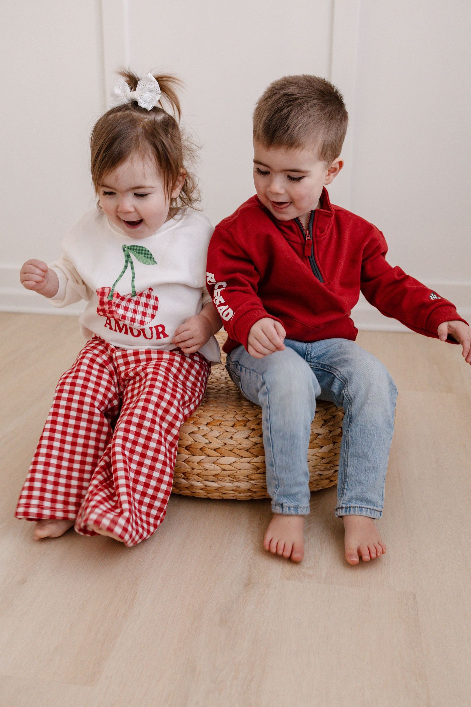Two children sit on woven basket wearing a red sweater and gingham pants laughing together during Lakeville mini session.