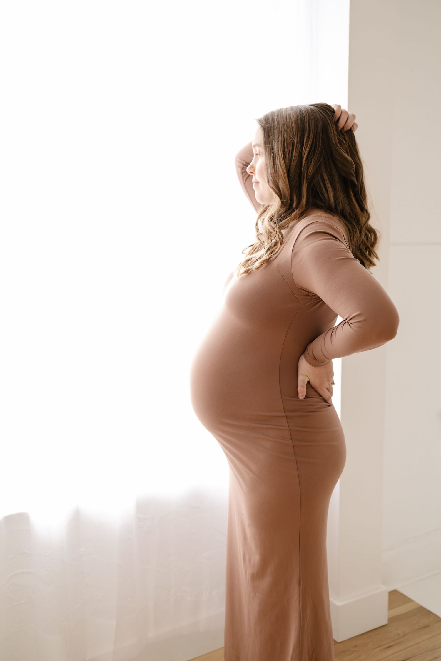 A pregnant woman in a brown dress stands in silhouette against a bright window in a minimalist room taken by Woodbury photographer.