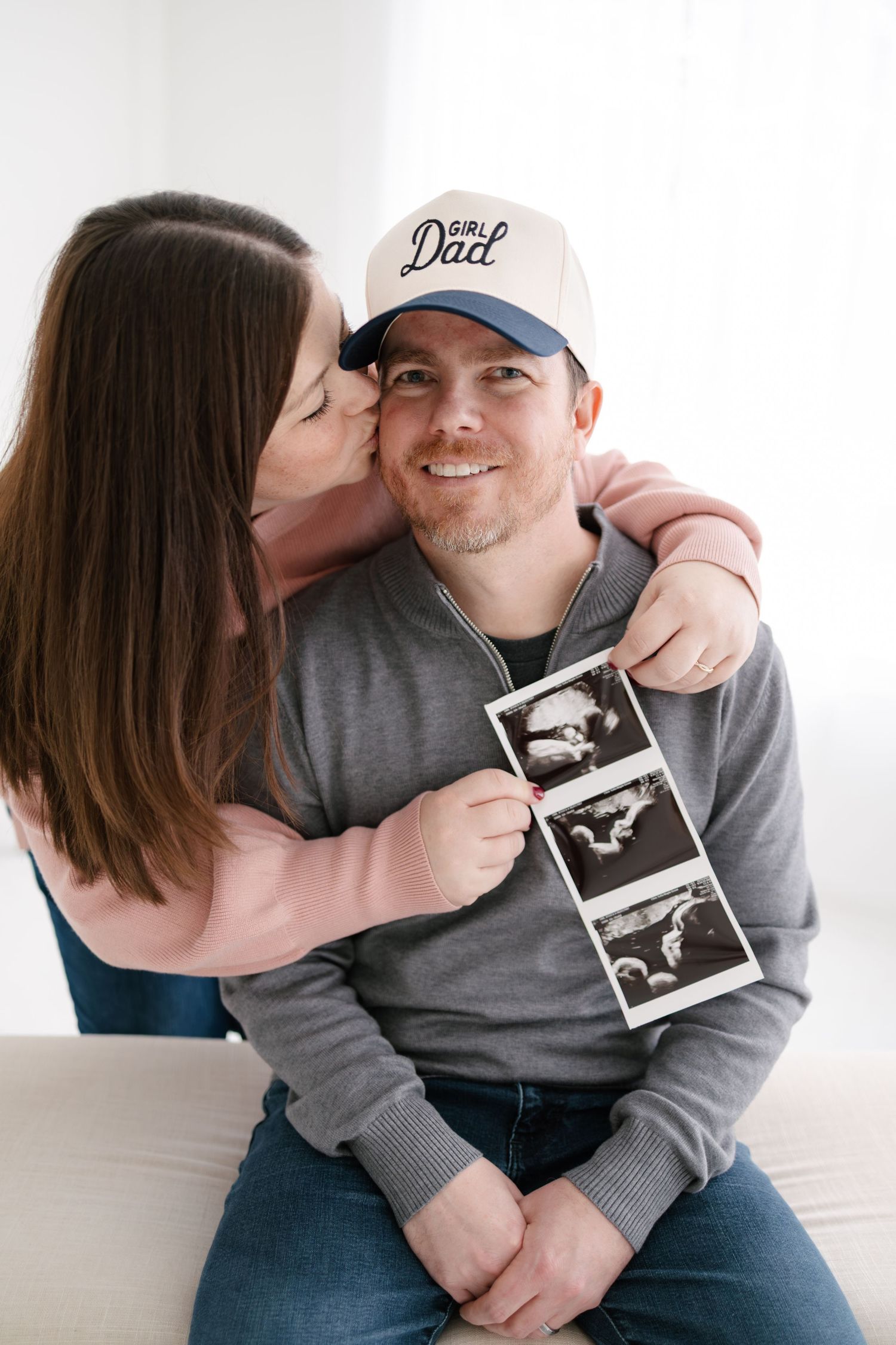 Happy couple holding ultrasound, man wearing a Girl Dad hat, woman kissing his cheek, announcing pregnancy with Woodbury photographer