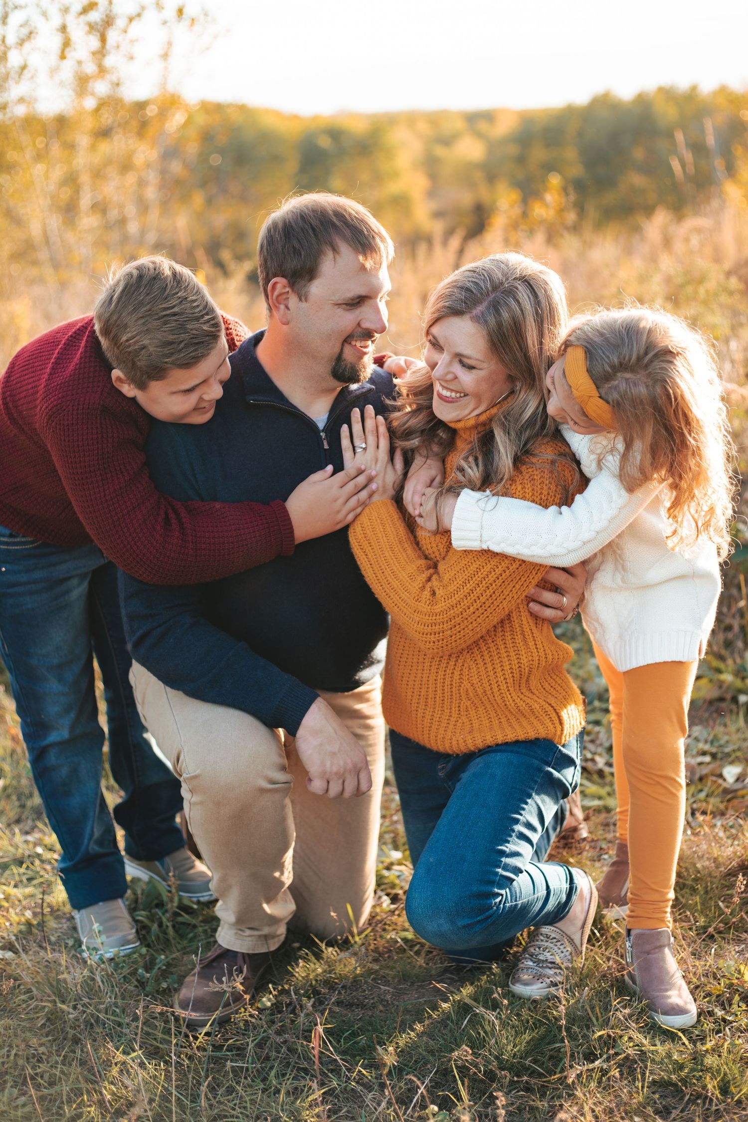 Happy family of four laughing together outdoors in a golden autumn field during a fall photo session taken by Woodbury family photographer.