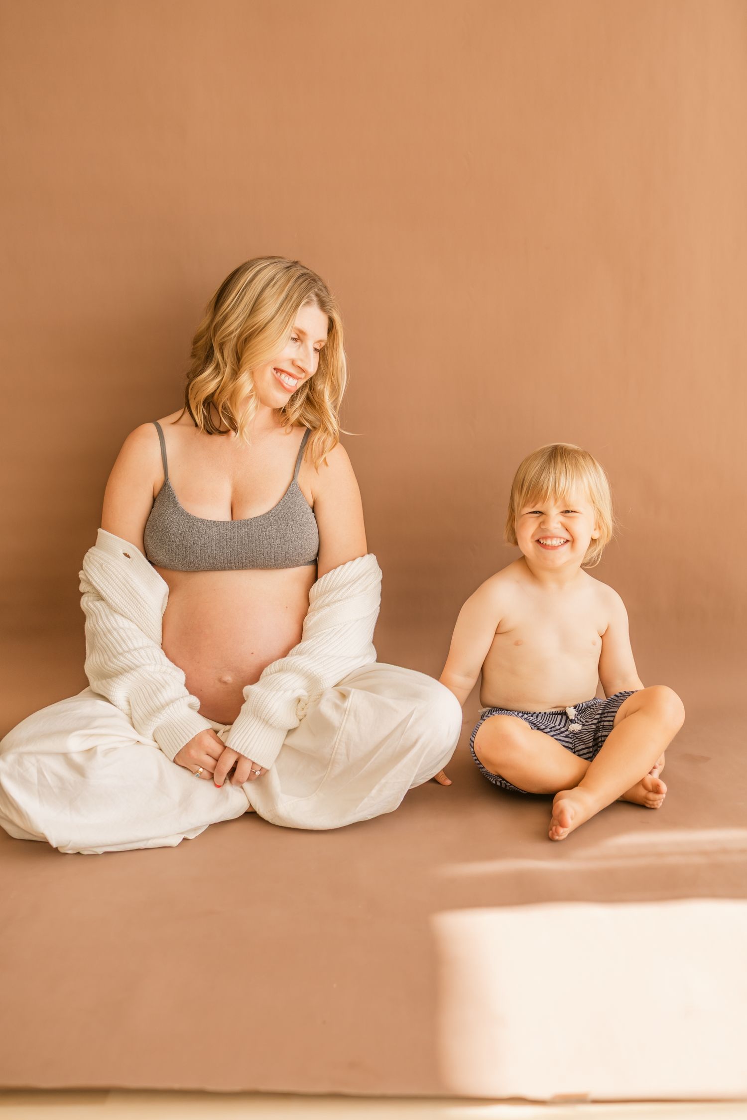 Pregnant woman in gray crop top sits with smiling toddler boy against warm beige background.
