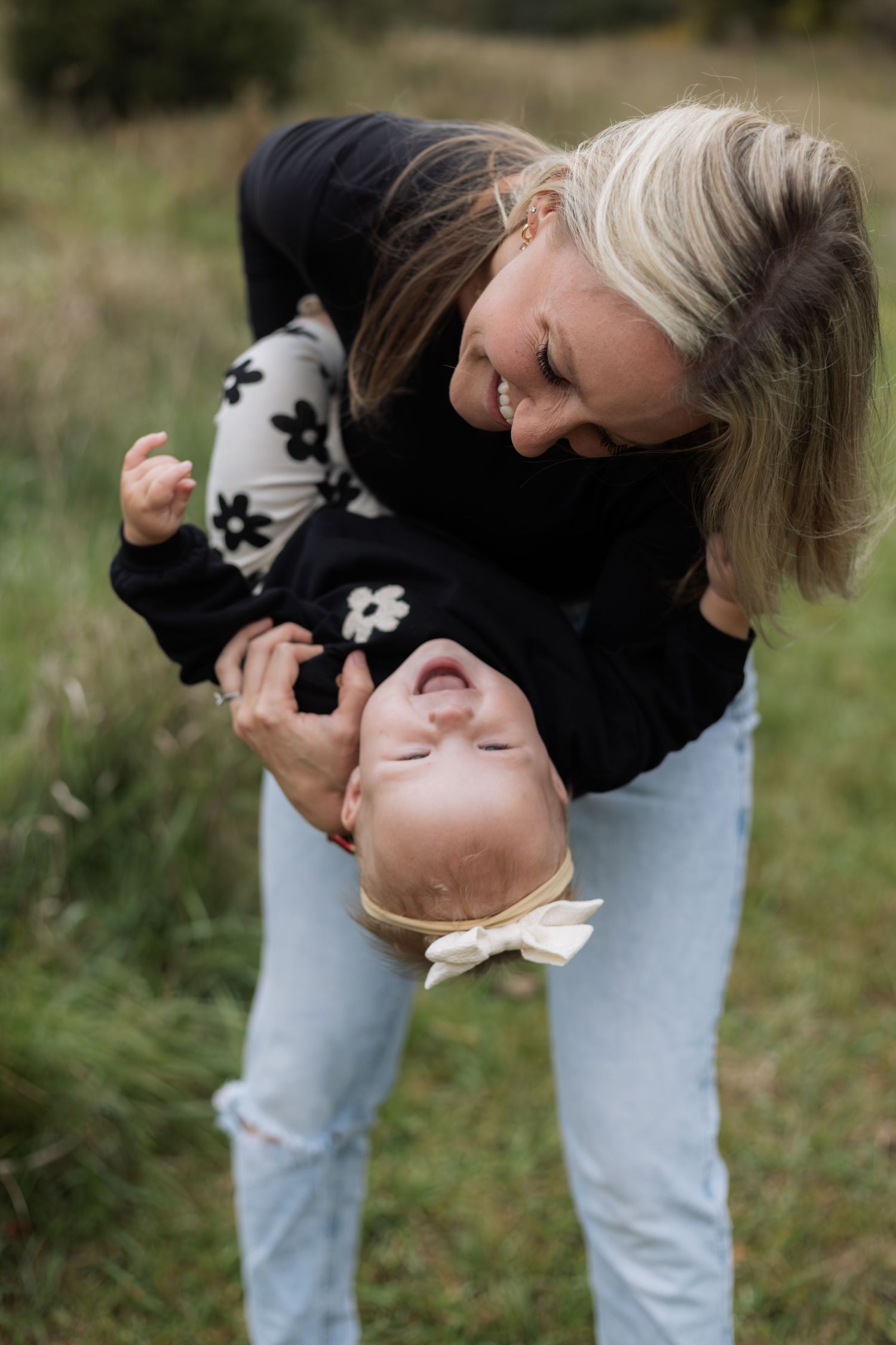A mother in light jeans and dark top bends down to play with a baby in the grass during Hudson fall mini session.
