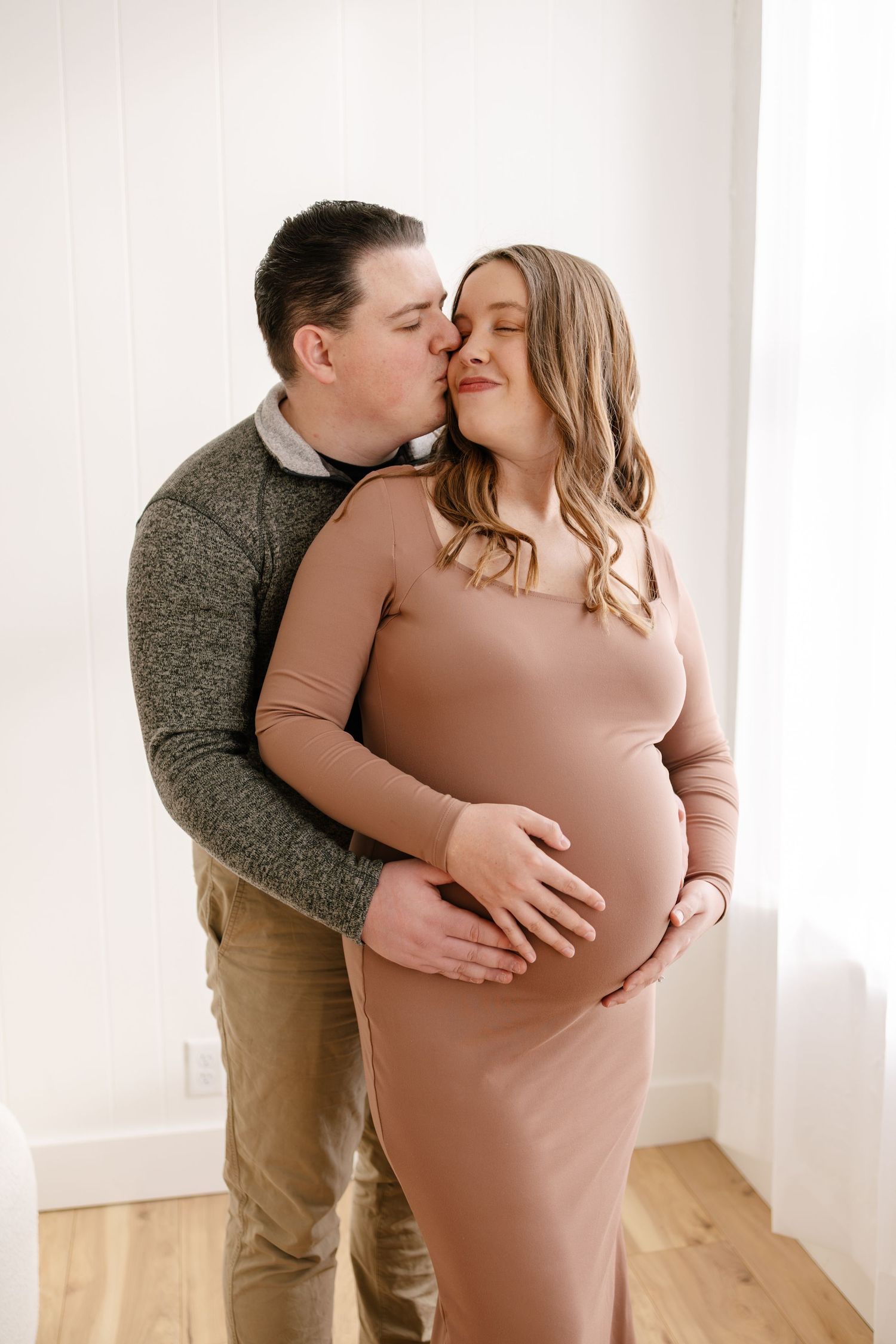 Couple sharing a tender maternity moment, man kissing pregnant woman's cheek while cradling her baby bump in a bright studio in Minneapolis