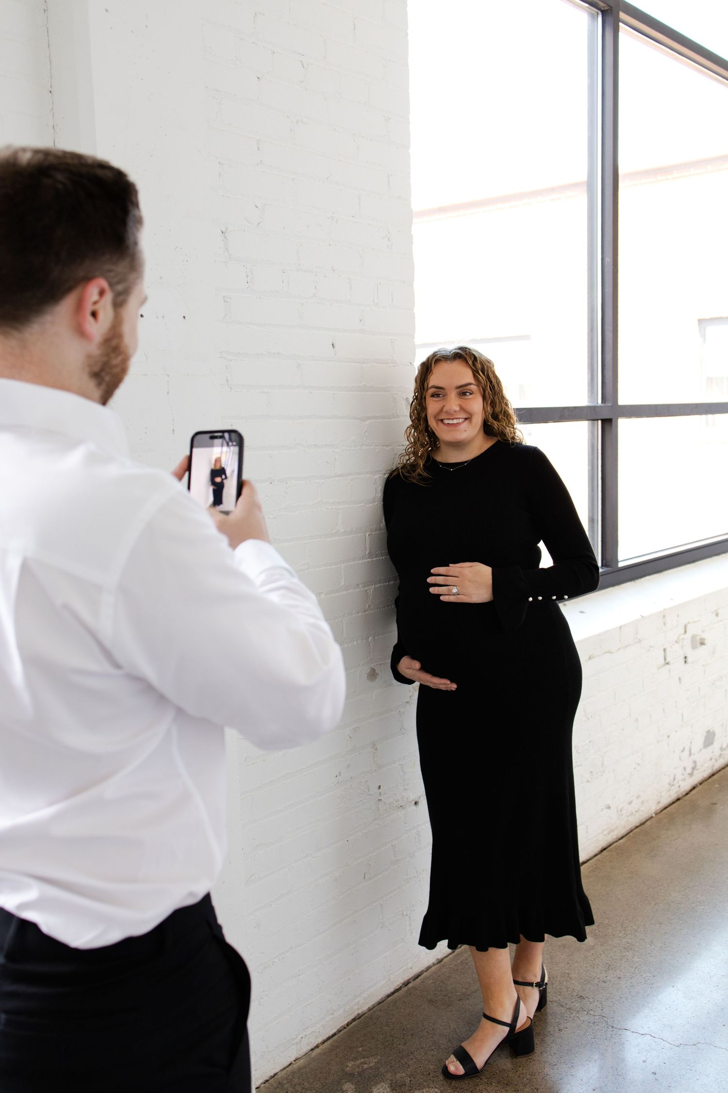A man photographs a smiling pregnant woman in a black dress against a white brick wall near a large window during maternity photo session