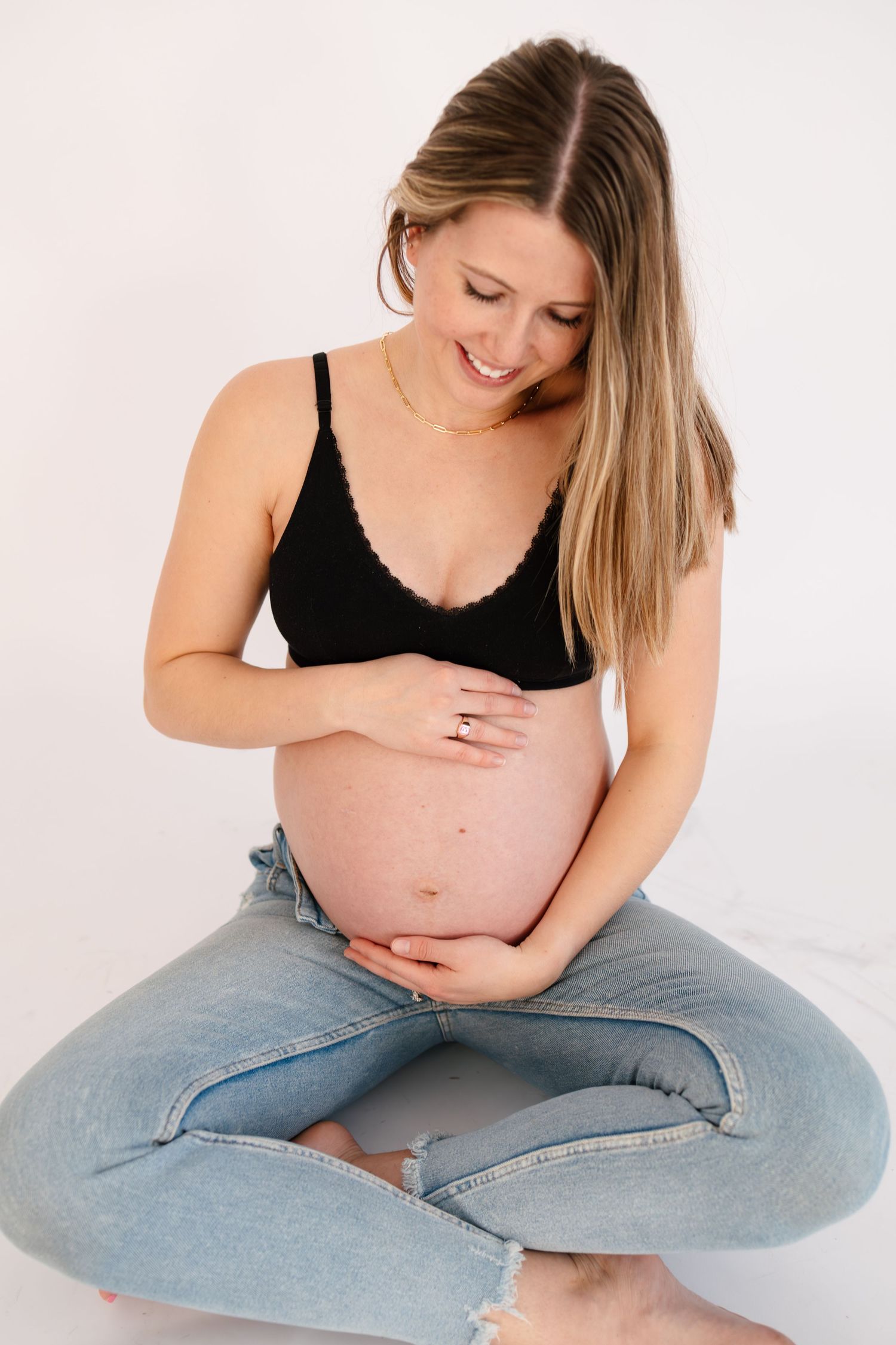 Smiling pregnant woman in black bralette and jeans sitting cross-legged, cradling her baby bump with both hands in Twin Cities studio